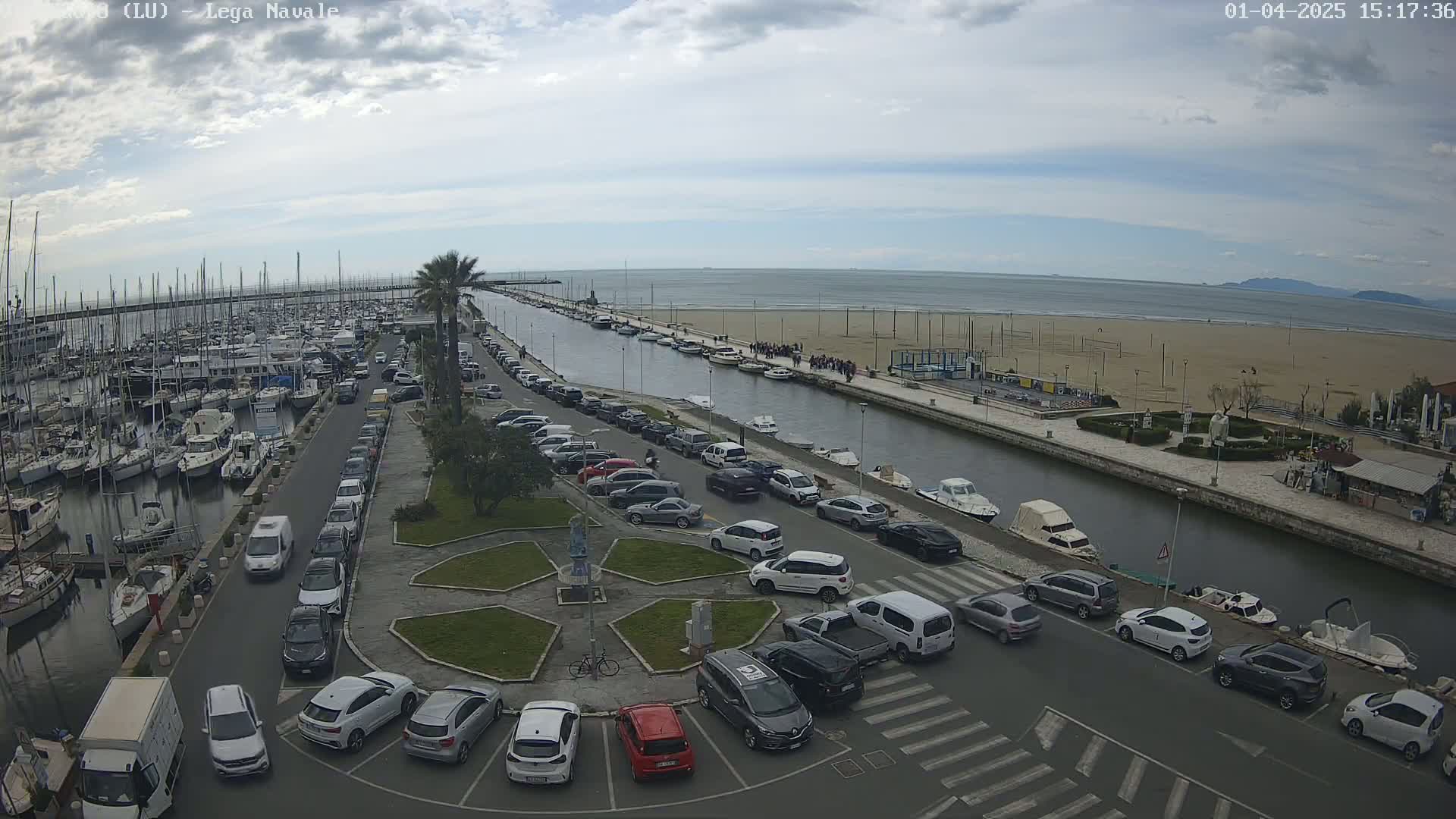 An overcast day shows a marina filled with boats, a canal with parked cars along its sides, a sandy beach, and the ocean in the distance.