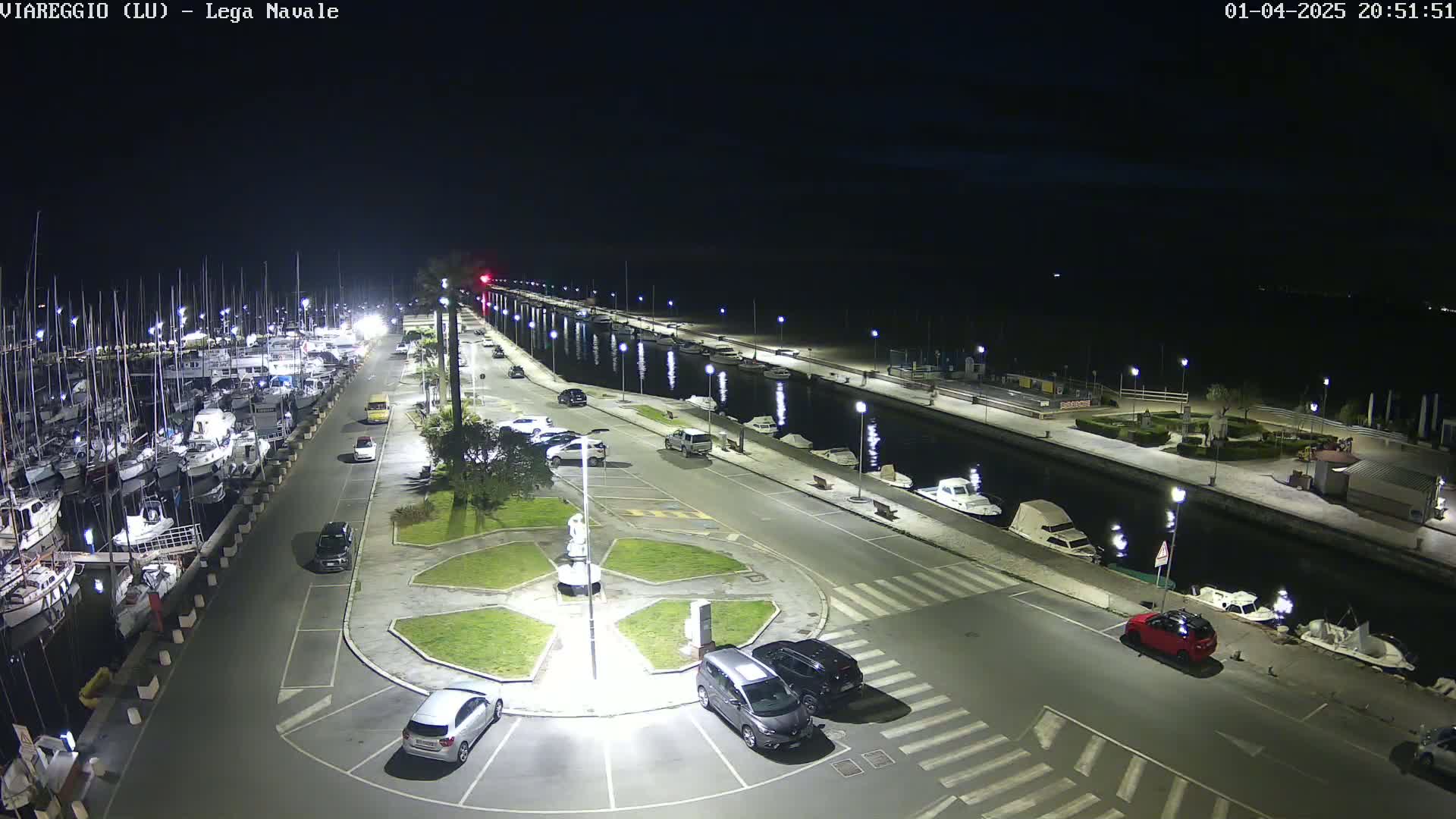 A nighttime, high-angle view of a marina with docked boats, cars parked in a lot, and a canal reflecting lights.