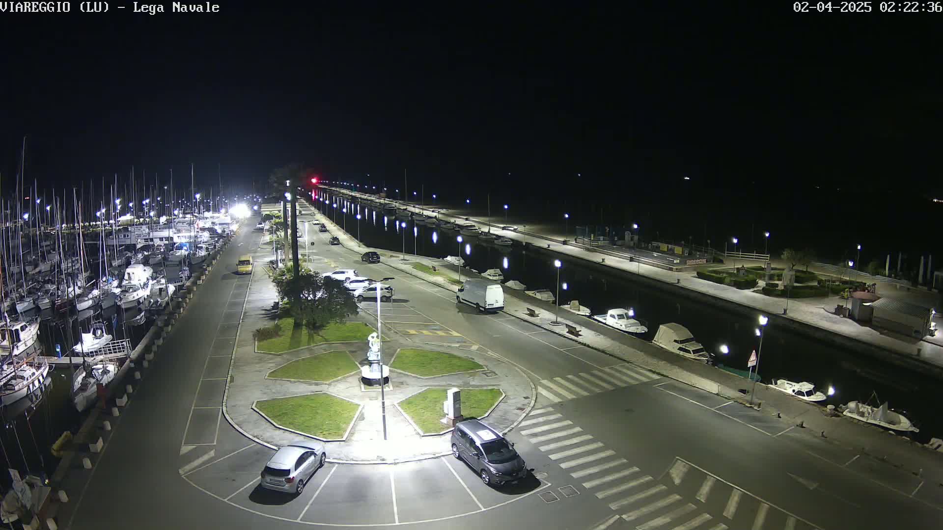 A nighttime, well-lit marina with numerous boats docked along a waterfront, and cars parked in a nearby lot, surrounding a small, landscaped traffic circle.
