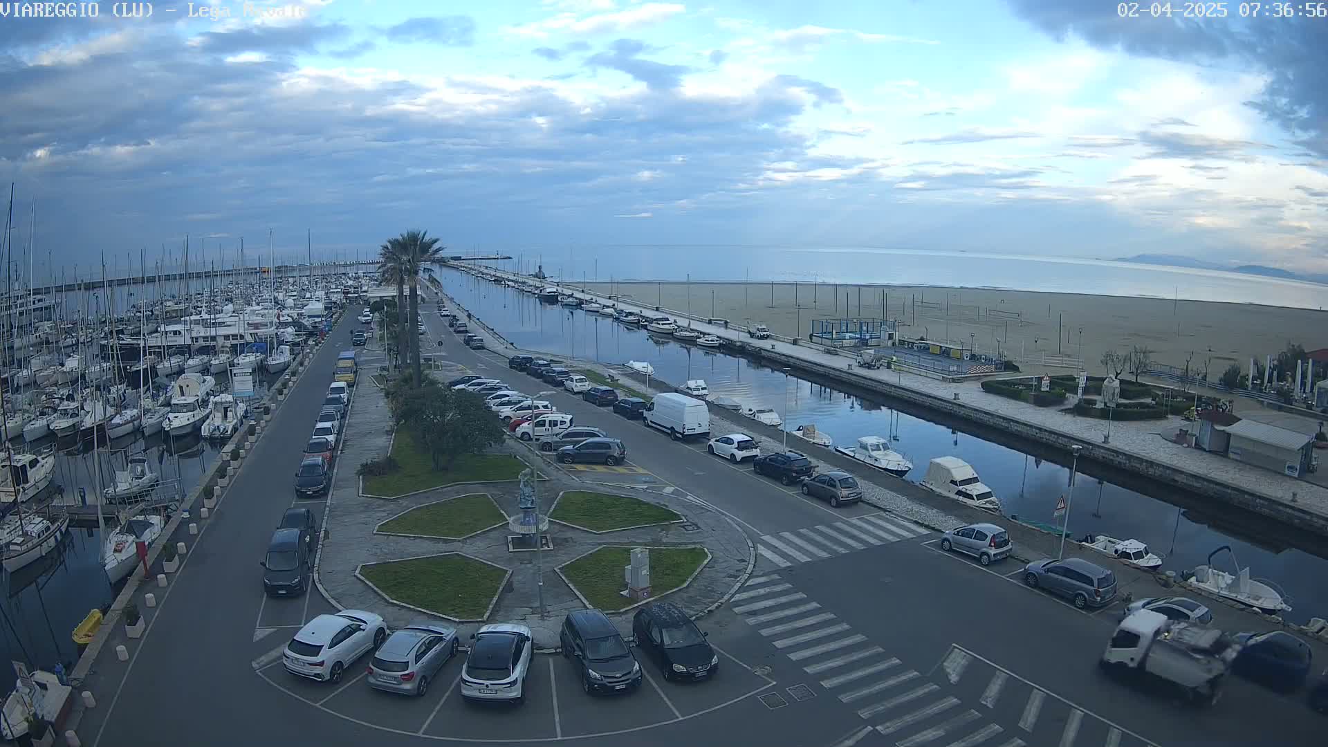 An overcast day shows a marina filled with boats, a canal lined with parked cars and a sandy beach beyond.