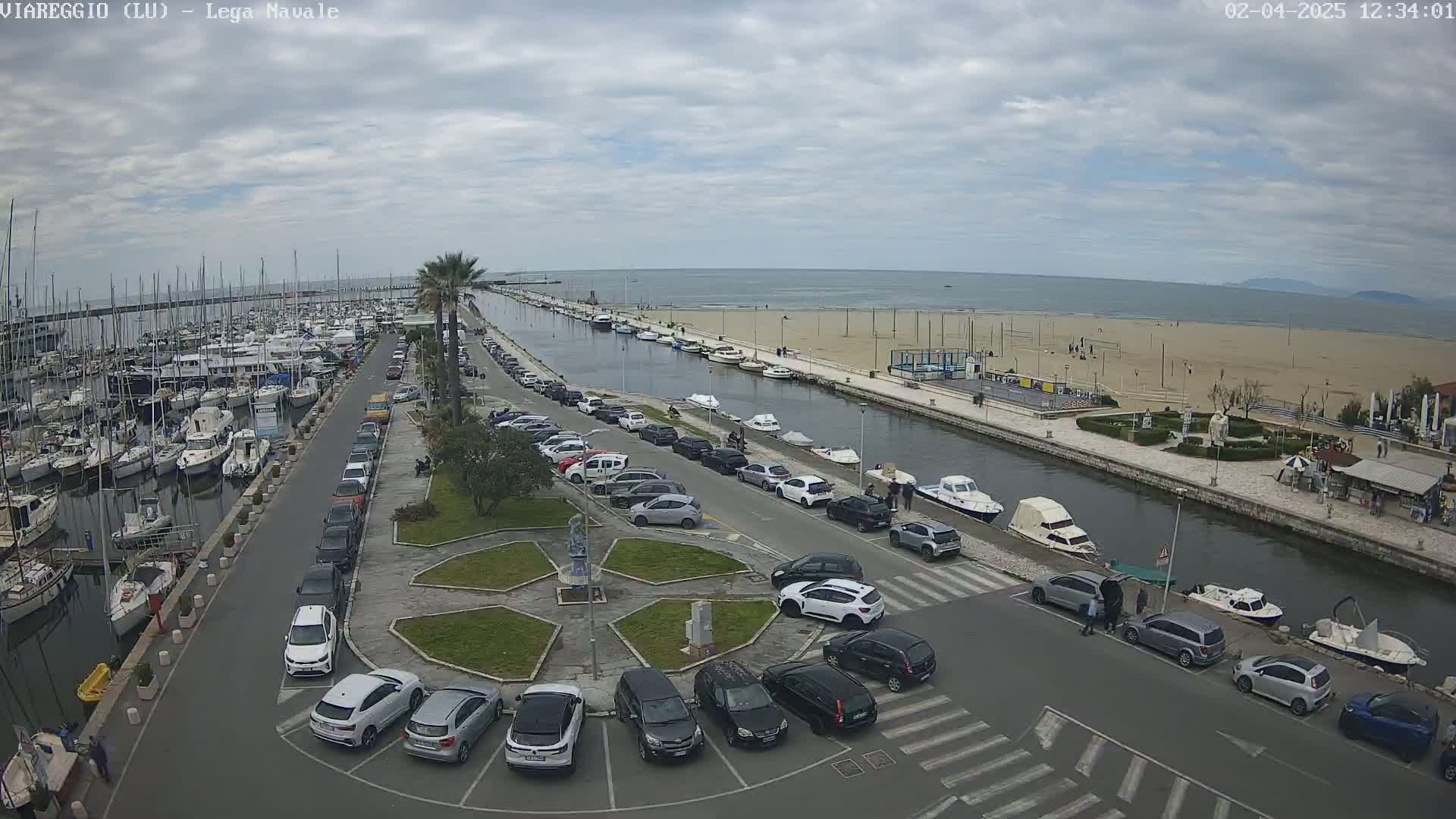 An aerial view shows a marina filled with boats, a canal lined with parked cars and small boats, a sandy beach, and an overcast sky.