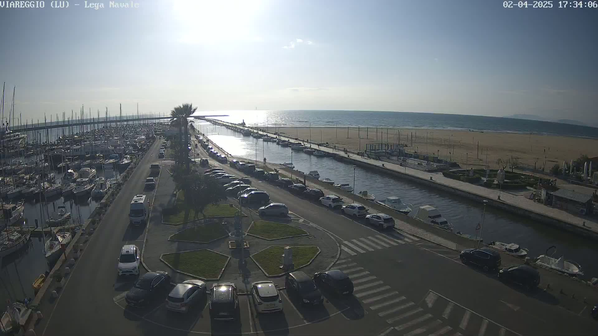 A sunny day at a harbor shows a marina filled with boats, a canal lined with parked cars, and a sandy beach beyond.