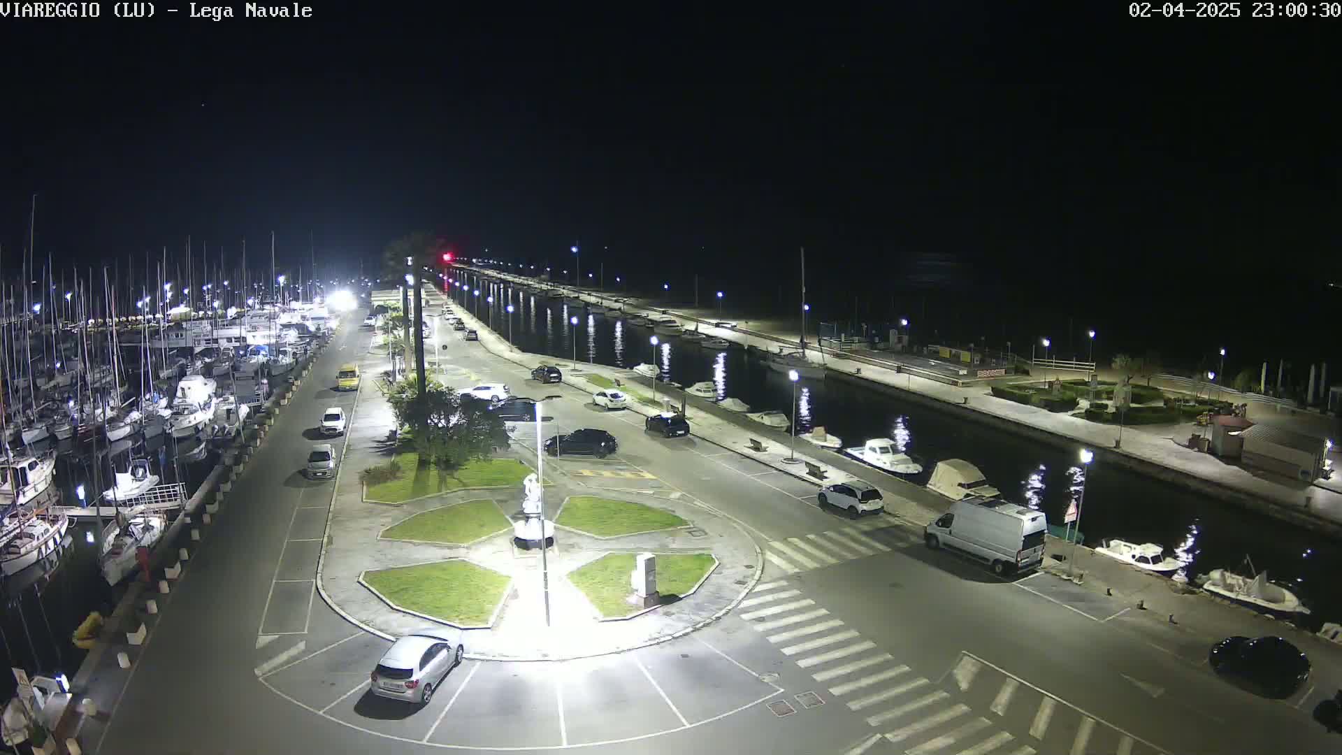 A nighttime aerial view of a marina with numerous boats docked, cars parked along a paved walkway, and a central roundabout.