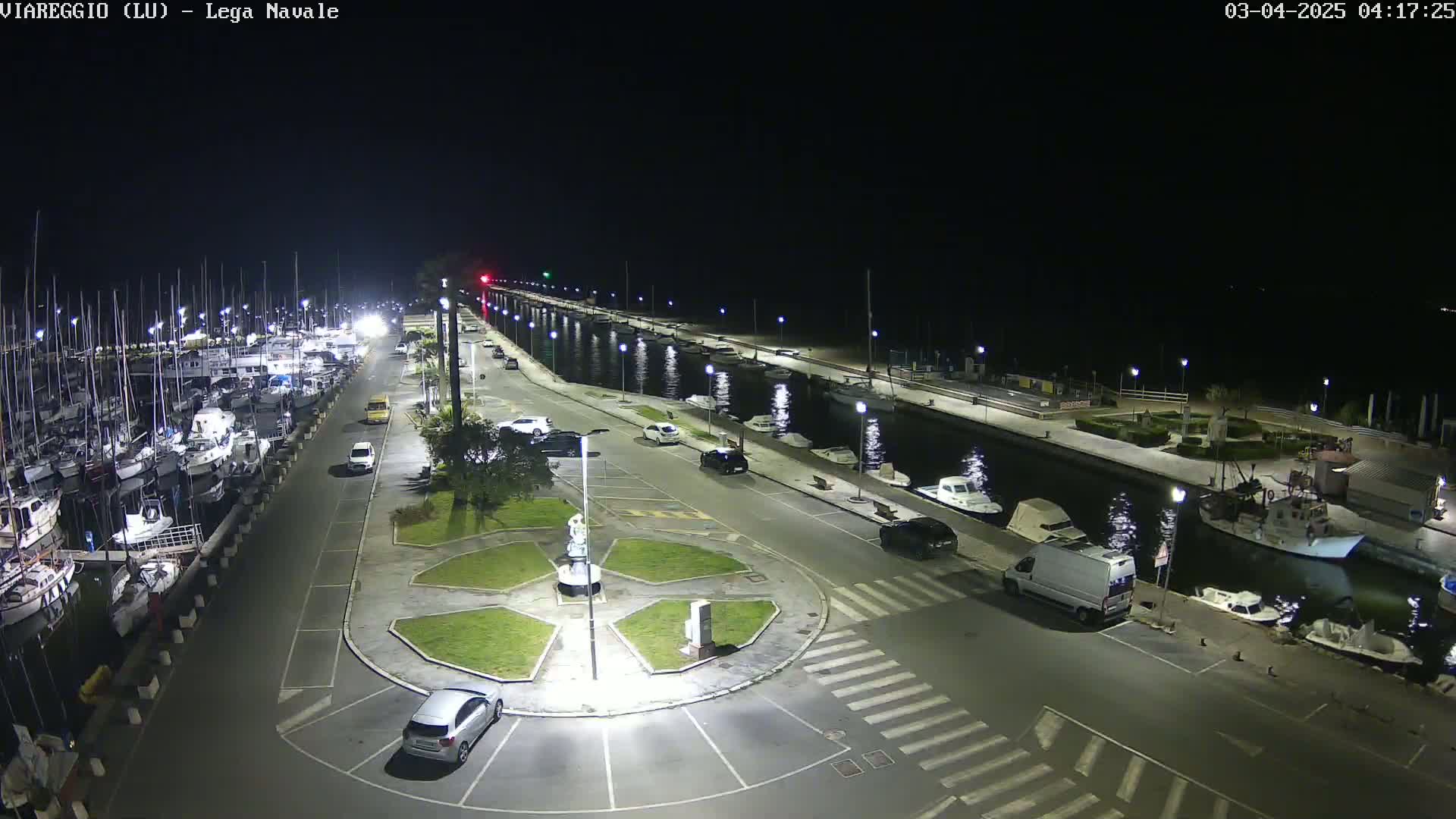 A nighttime, high-angle view of a marina with docked boats, cars parked along a waterfront road, and a small roundabout in the foreground.