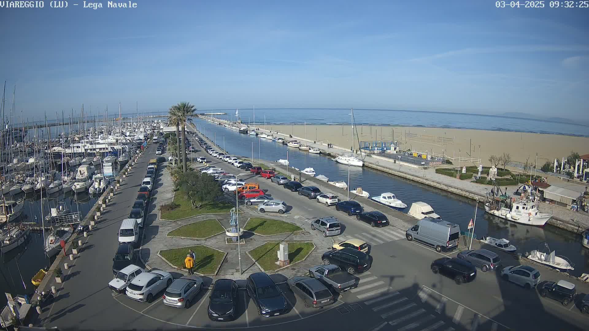 A marina filled with boats is adjacent to a sandy beach and calm ocean under a clear blue sky, with a parking lot and canal separating them.