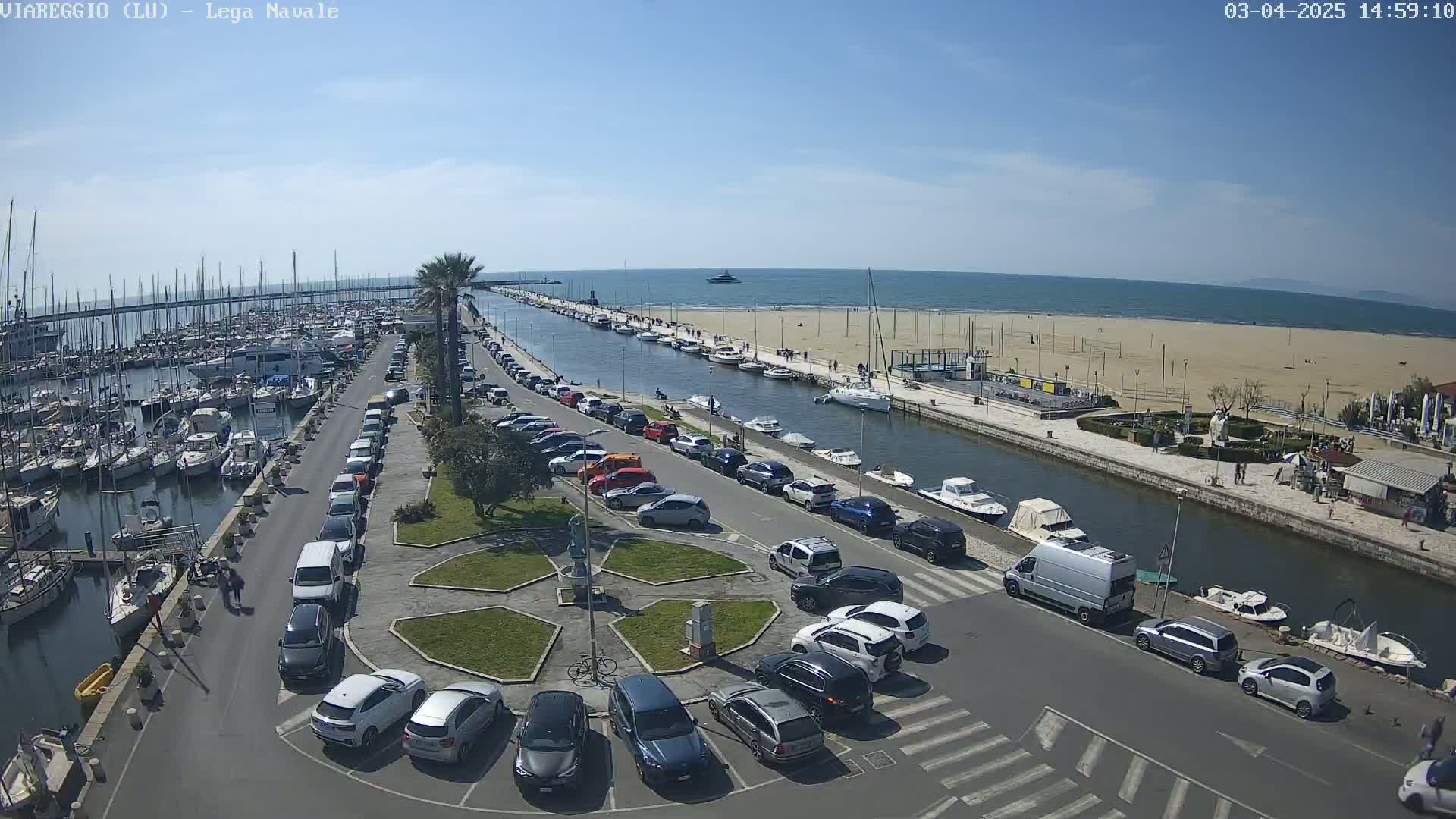 A marina filled with boats is adjacent to a sandy beach and calm ocean under a clear blue sky, with cars parked along a canal separating the two.