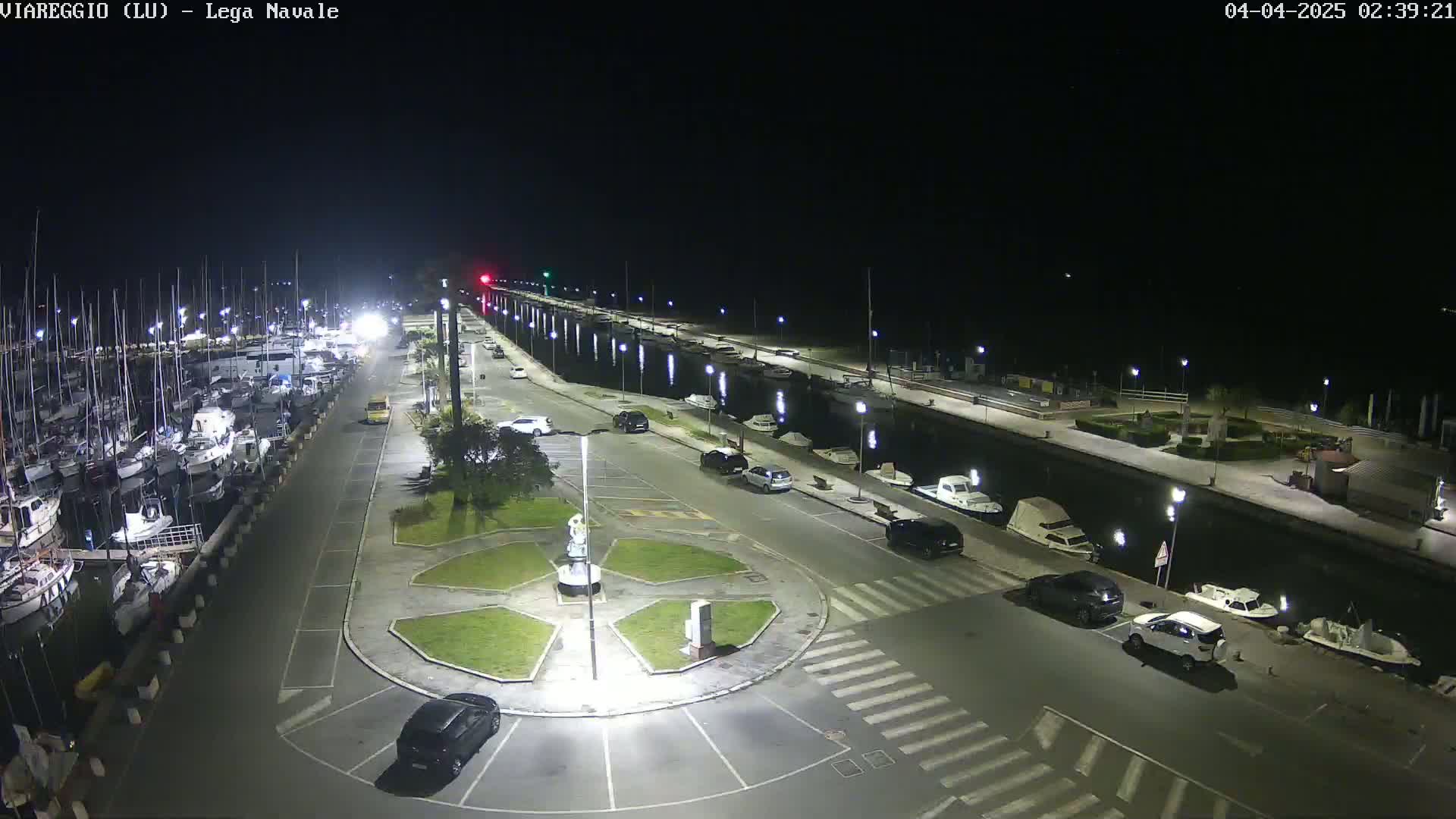 A nighttime aerial view of a marina with docked boats, a paved area with parked cars, and a small roundabout in the center.