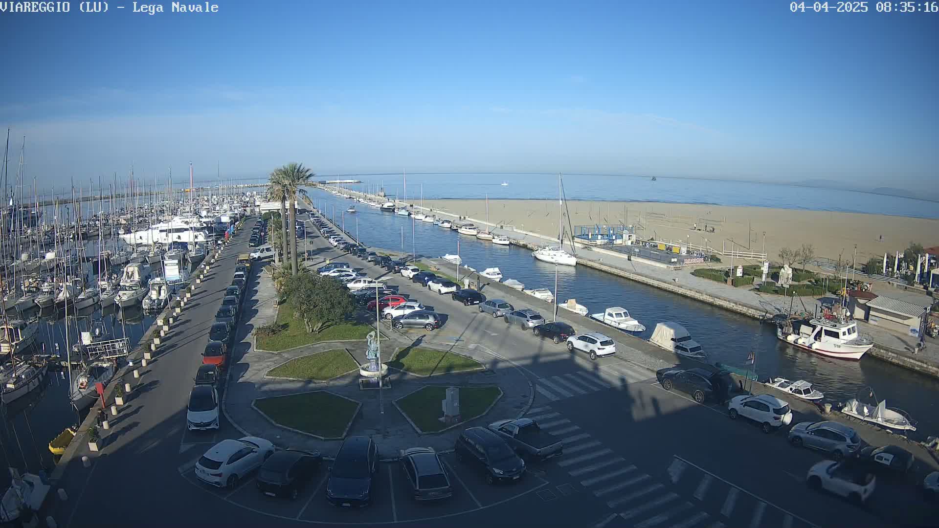 A marina filled with boats and yachts is adjacent to a sandy beach and canal, with cars parked along the waterfront under a clear blue sky.