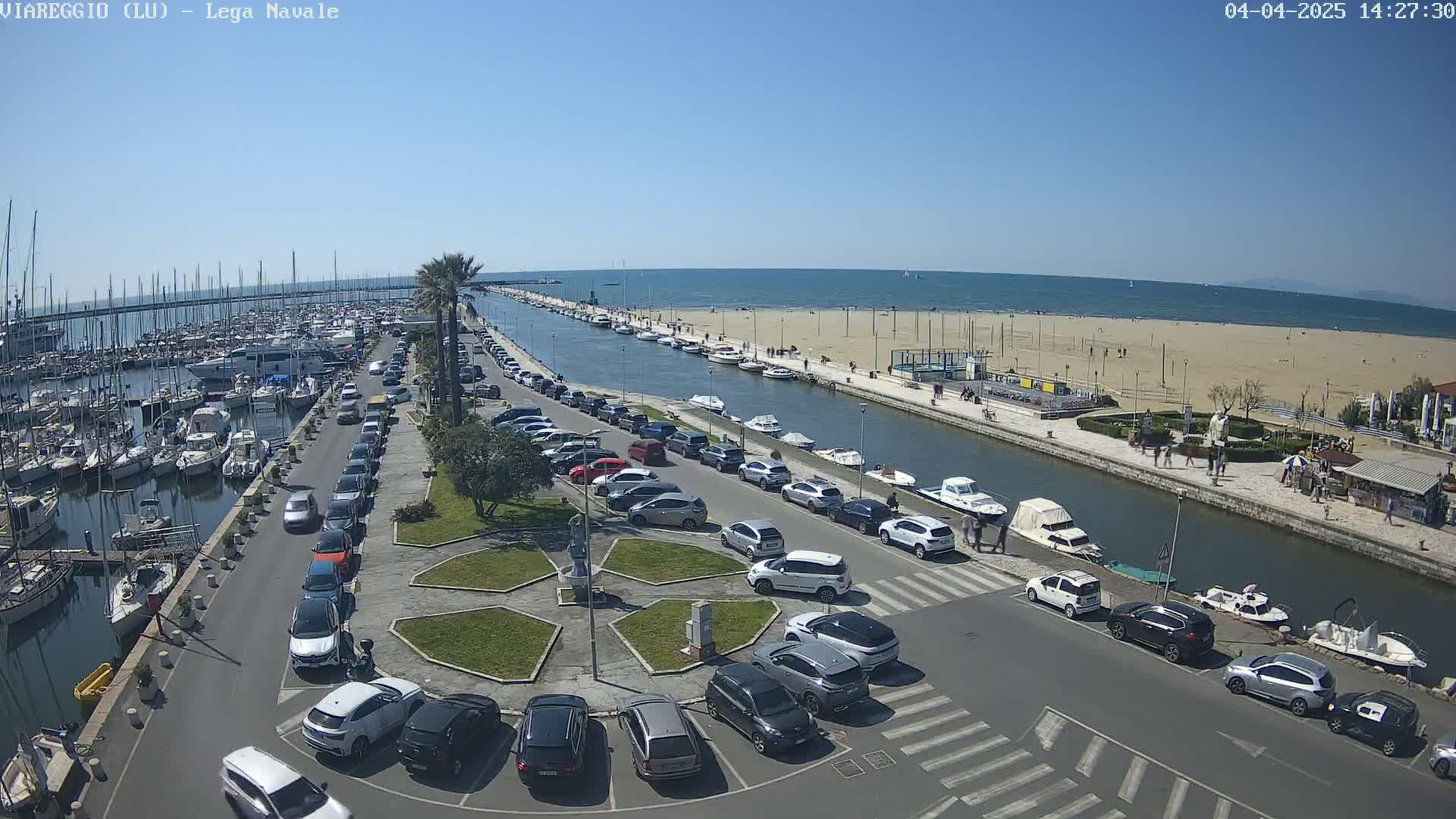 A marina filled with boats and surrounded by cars parked along a canal leading to a sandy beach on a sunny day.