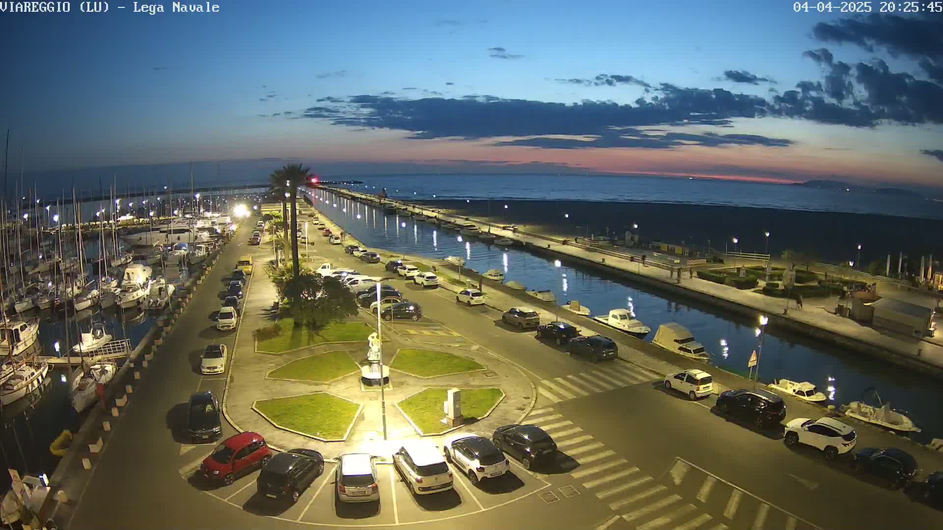 An aerial view of a marina at dusk, with numerous boats docked and cars parked along a canal leading to a beach under a partly cloudy sky.