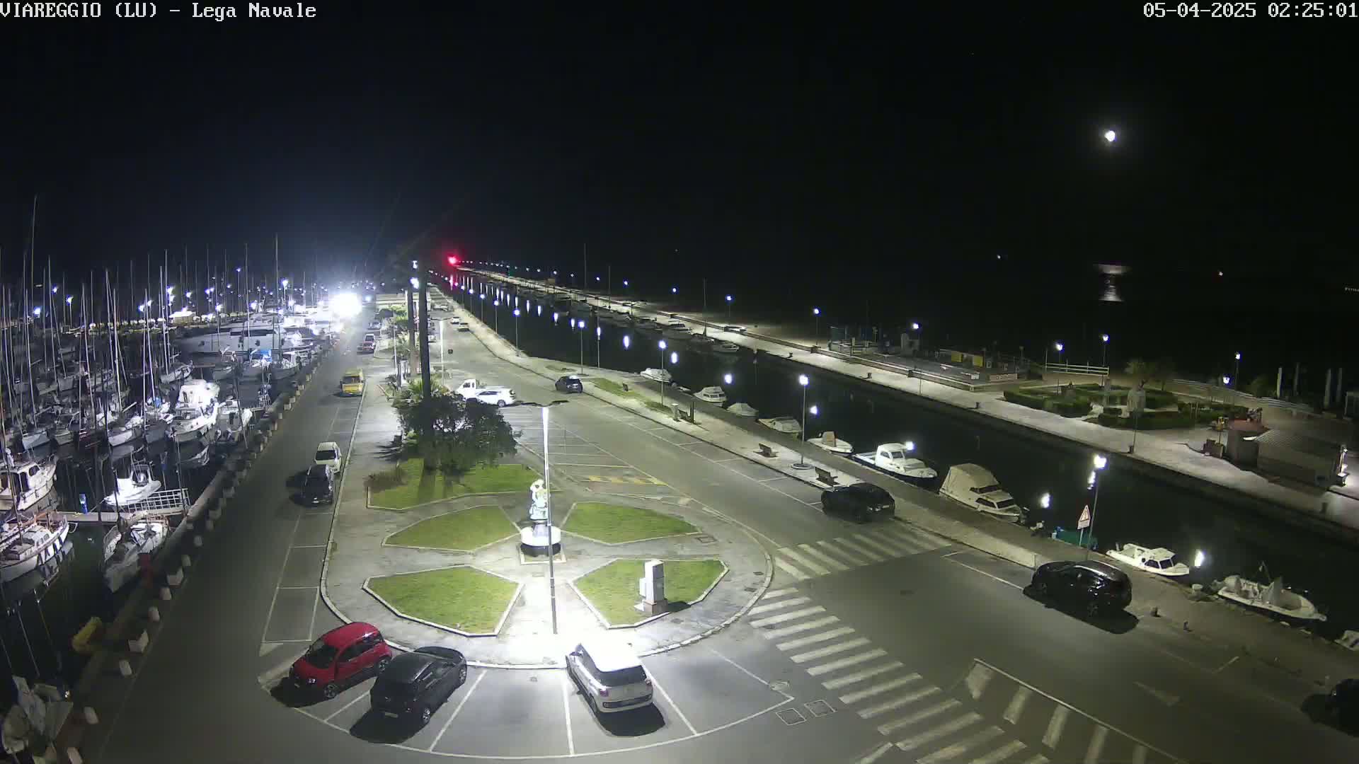A nighttime, well-lit marina with numerous boats docked, cars parked along a paved roadway, and a small central plaza.