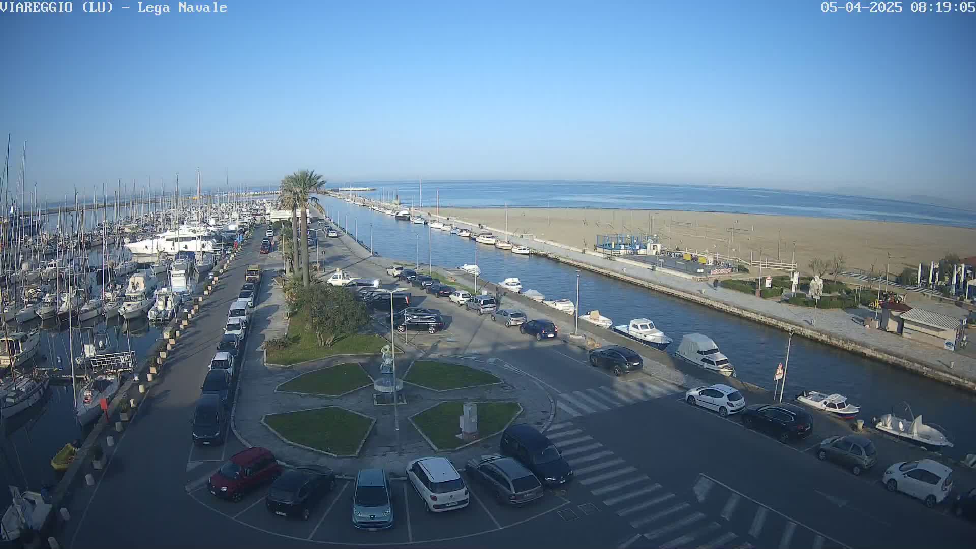 A marina filled with boats is adjacent to a sandy beach and calm ocean under a clear blue sky.