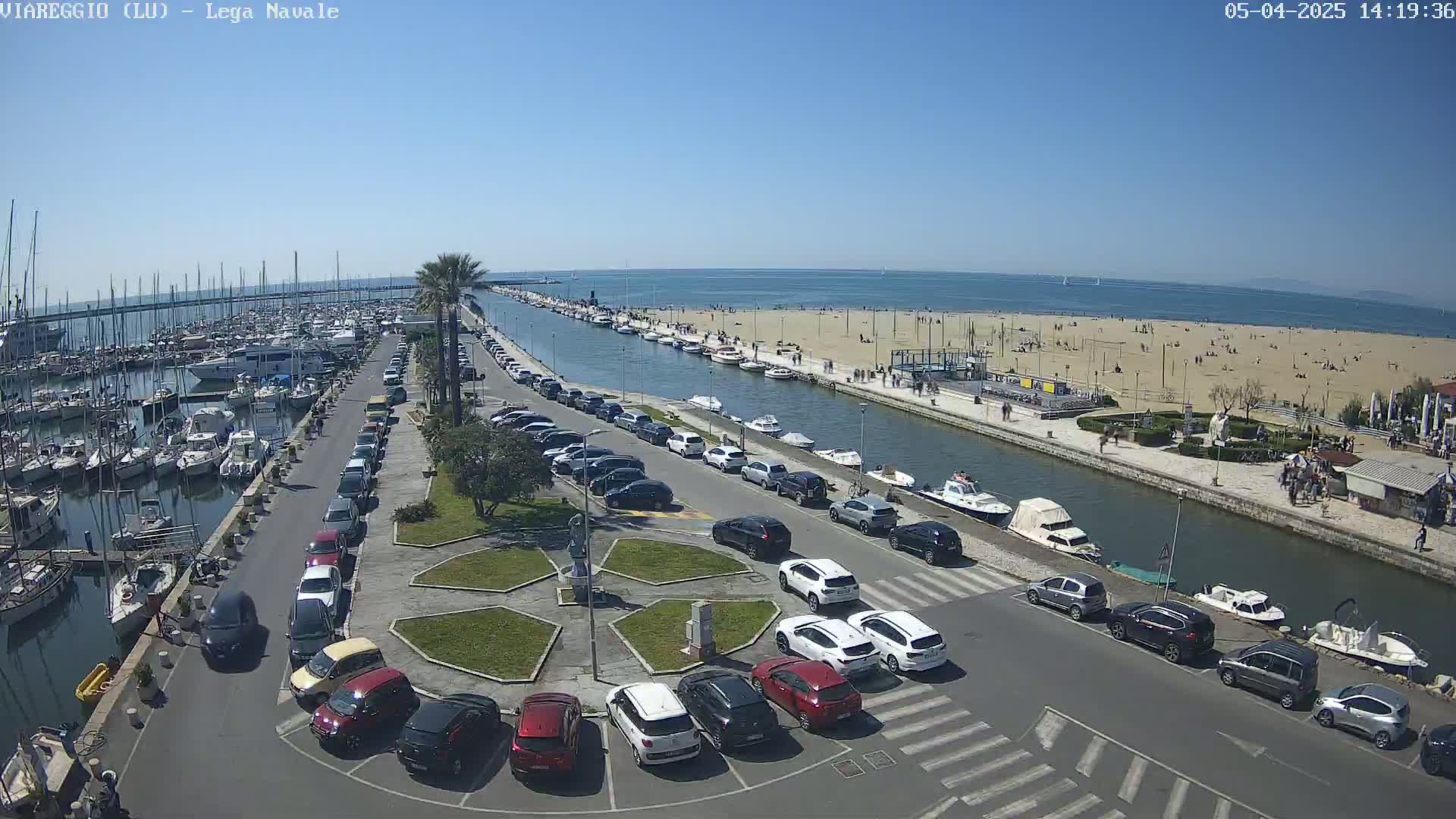 A marina filled with boats is adjacent to a canal and a sandy beach with people sunbathing under a clear blue sky.