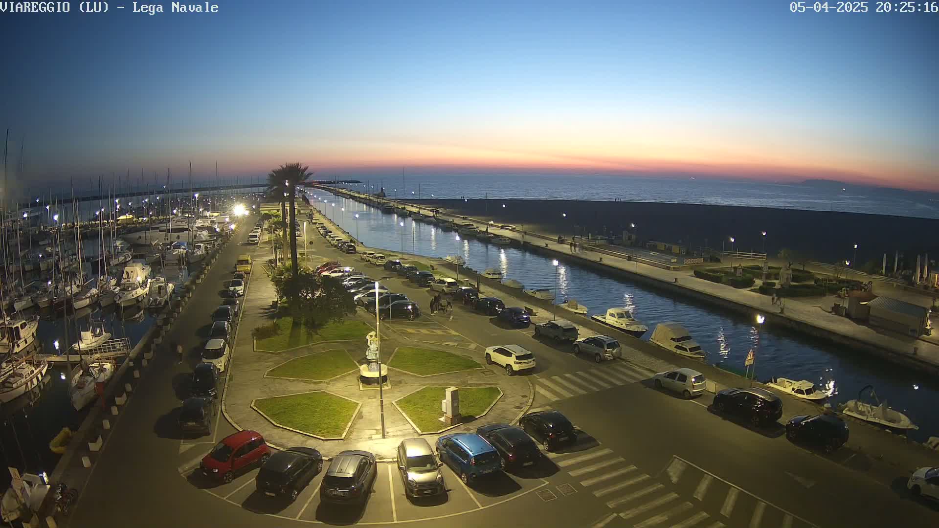 An aerial view at dusk shows a marina filled with boats, a canal lined with parked cars, and a beach in the background under a twilight sky.