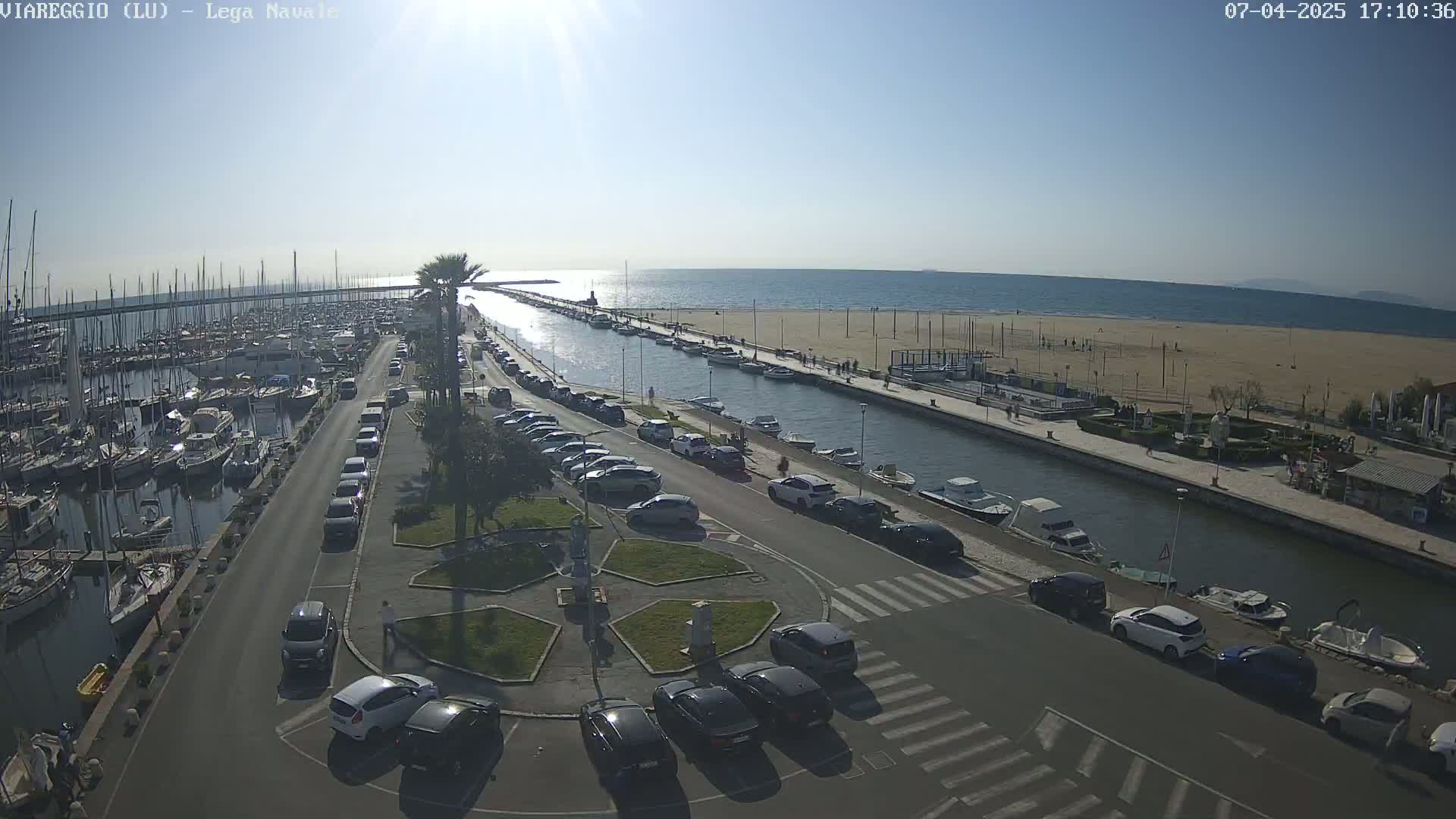 An aerial view shows a marina filled with boats, a canal leading to a sandy beach under sunny skies, and a parking lot filled with cars.