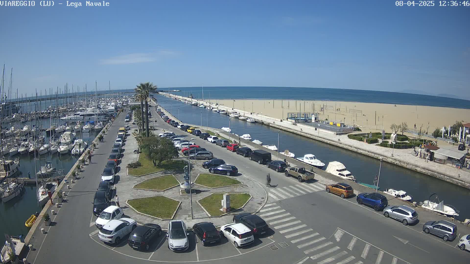A marina filled with boats, a canal, a sandy beach, and a parking lot with cars are visible on a sunny day.