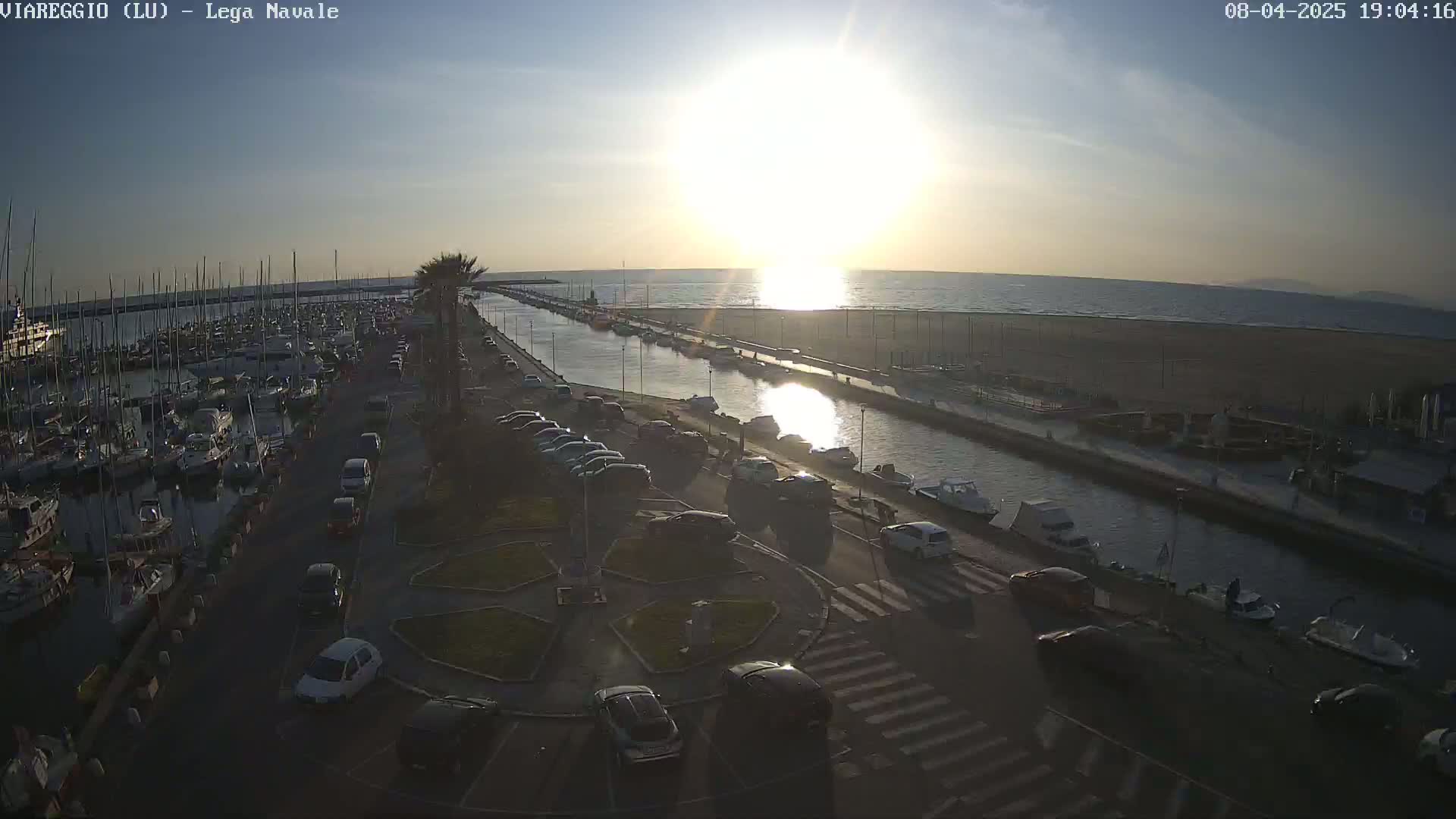 A marina filled with boats is shown at sunset next to a canal and sandy beach with cars parked along the waterfront.