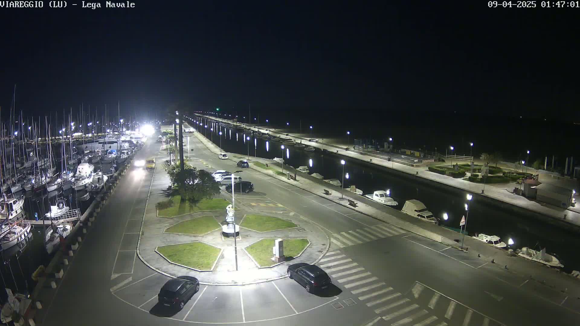 A nighttime view of a marina with docked boats and cars parked along a paved area next to a canal.