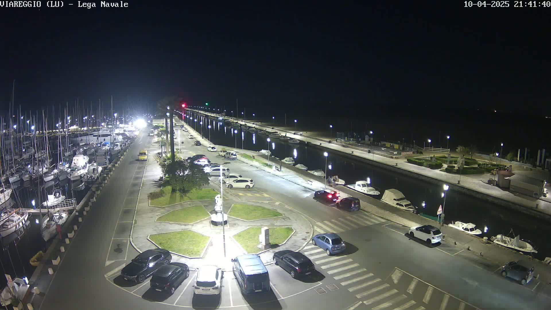 A nighttime aerial view of a marina with numerous boats docked, a parking lot with several cars, and a canal separating the parking lot from a waterfront promenade.