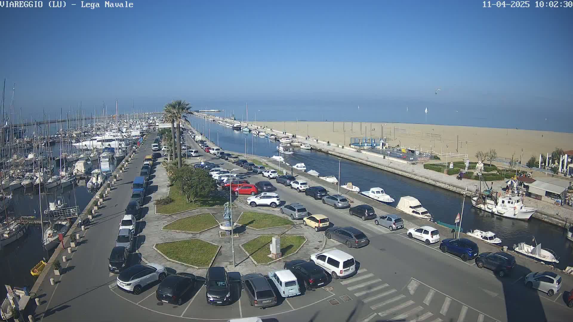 A marina filled with boats and surrounded by a parking lot, a canal, and a sandy beach under a clear blue sky.