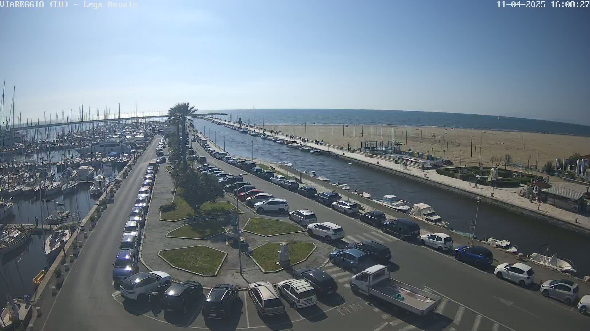 A marina filled with boats is next to a canal that leads to a sandy beach on a sunny day.