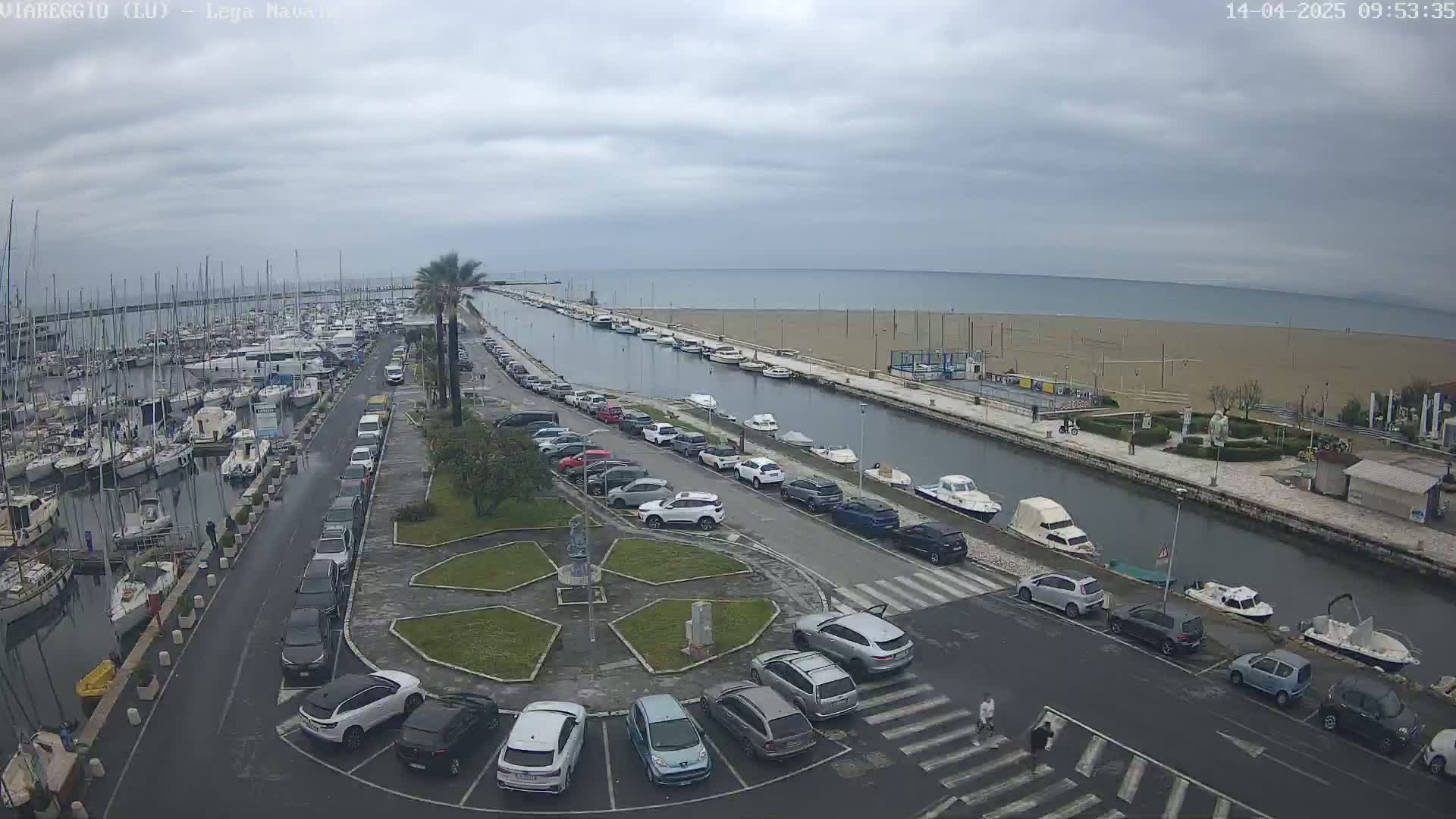 An overcast day reveals a marina filled with boats, a canal lined with parked cars, and a sandy beach beyond.