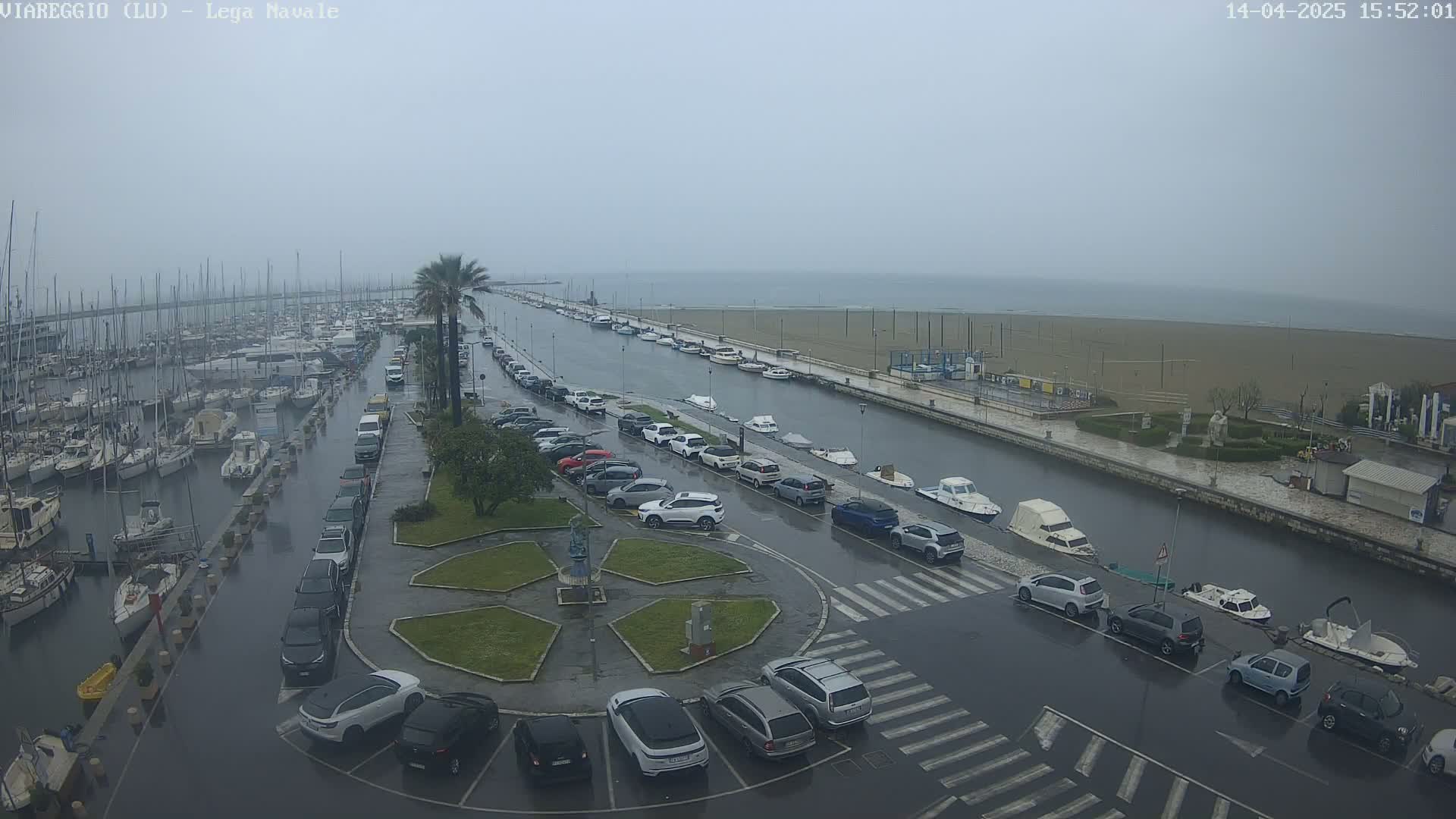 A rain-soaked marina and waterfront parking lot are shown, with numerous cars parked alongside a canal filled with boats and a sandy beach visible in the distance.