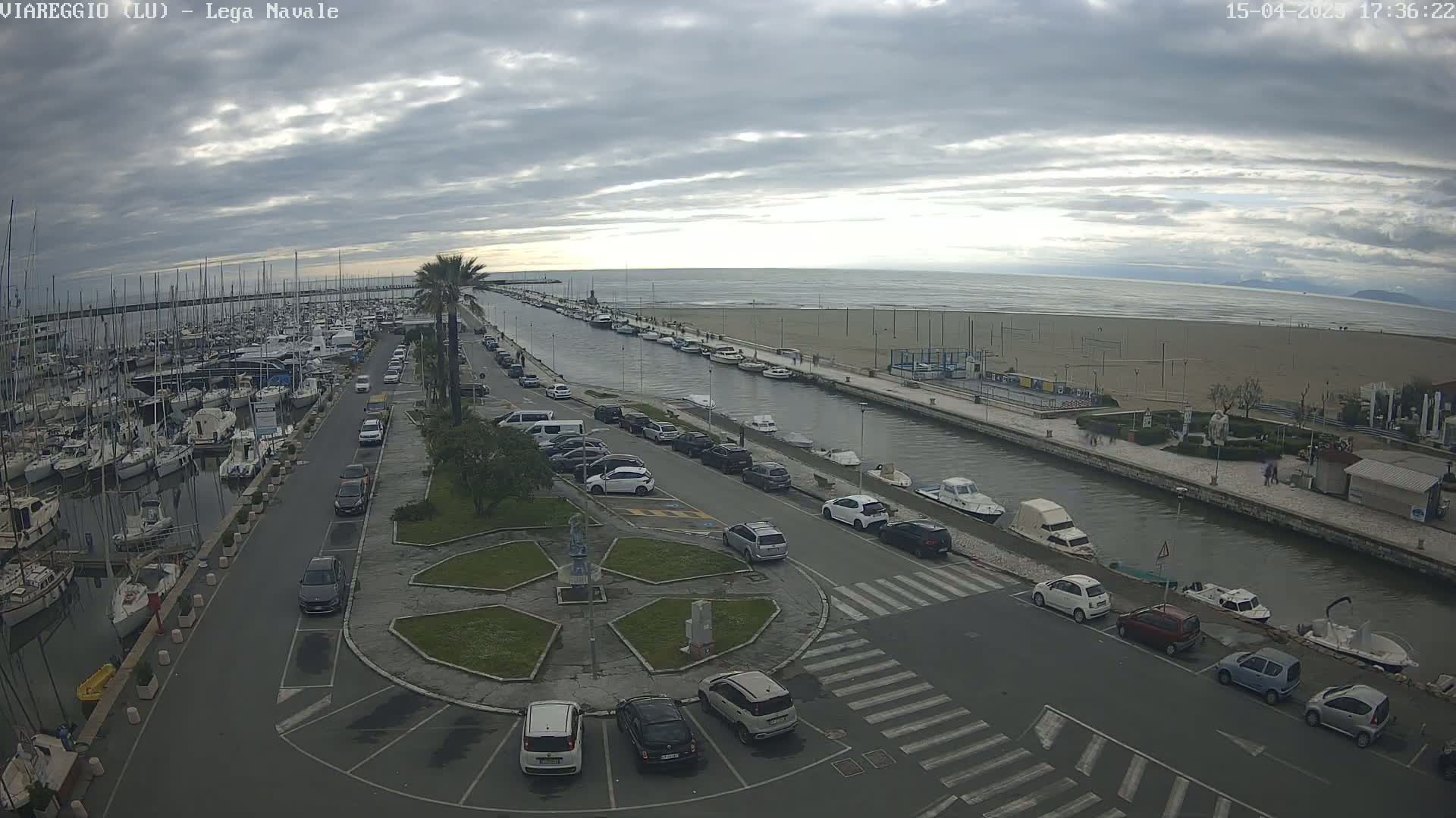 An overcast day shows a marina filled with boats, a canal with moored boats, and a parking lot next to a sandy beach.