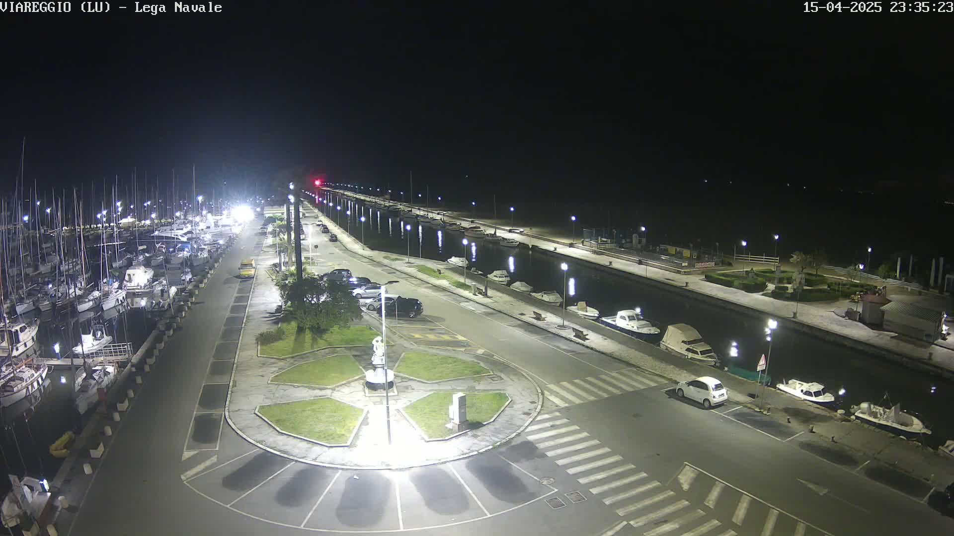 A nighttime aerial view of a marina with boats docked, a roadway with cars parked, and a small roundabout in the center, all brightly lit under clear skies.
