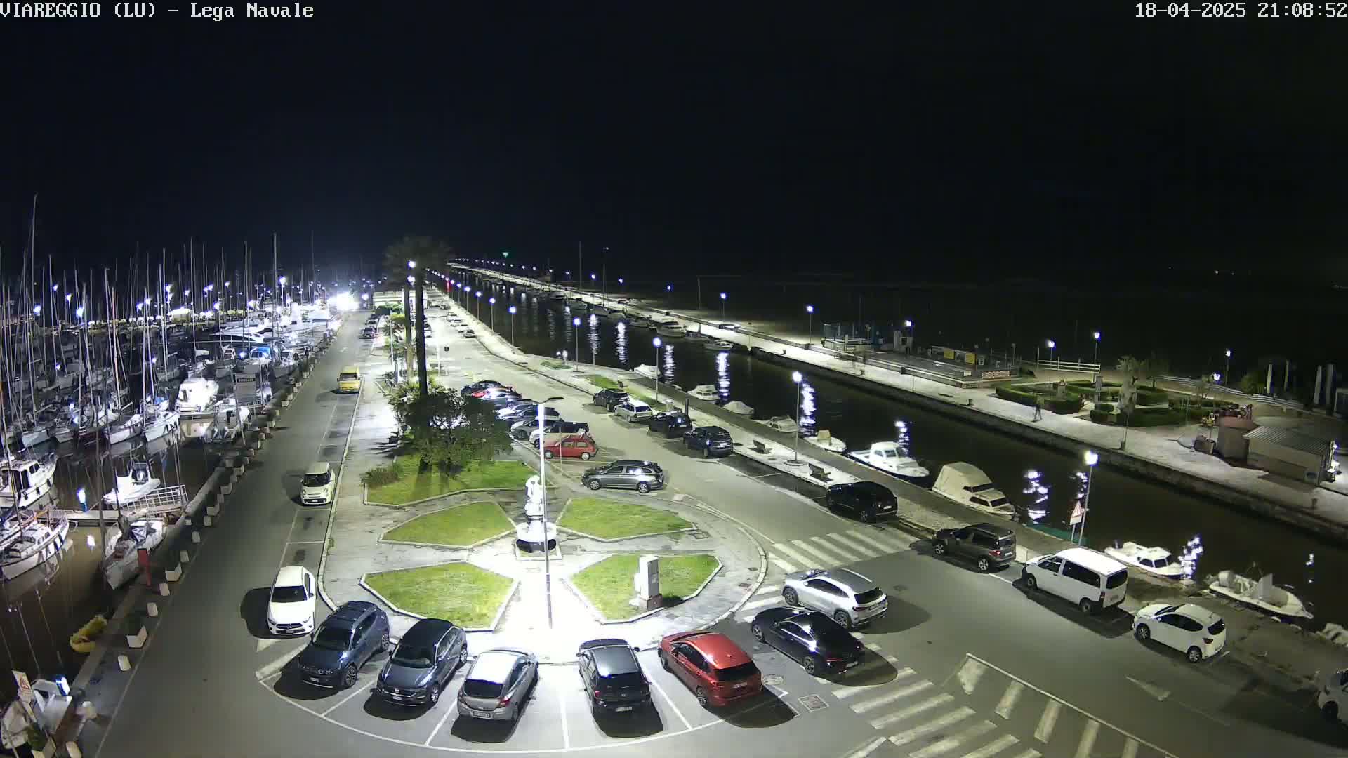 A nighttime aerial view of a marina with numerous boats docked, a canal, and a parking lot filled with cars.