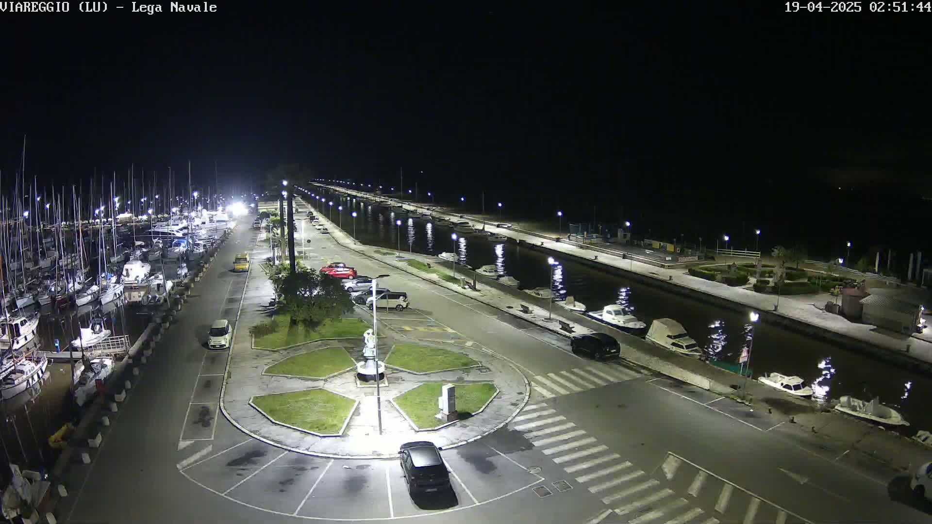 A nighttime aerial view of a marina with numerous boats docked, cars parked in a lot beside a canal, and a small roundabout in the foreground.