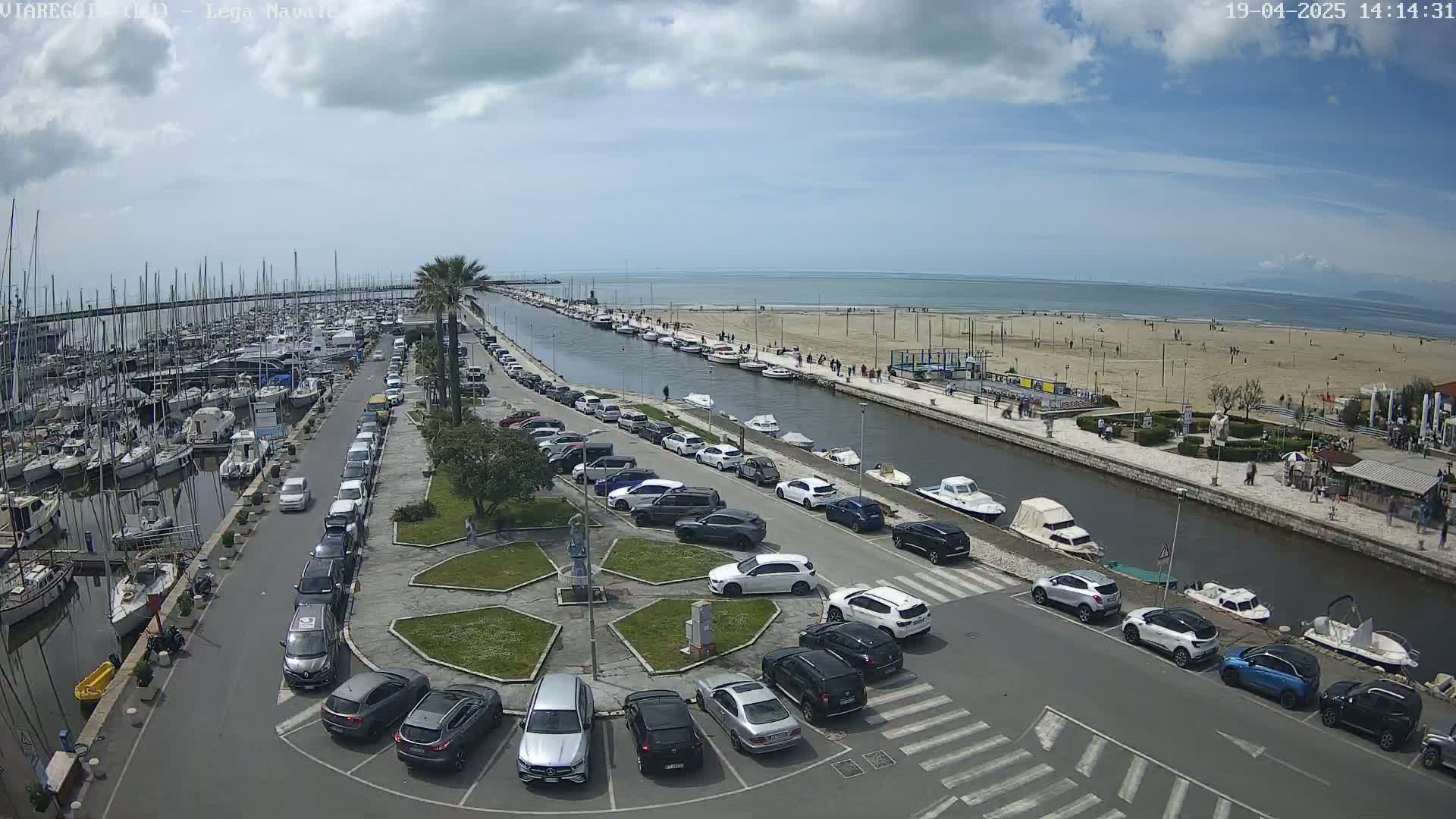 A marina filled with boats next to a canal lined with parked cars and a sandy beach under a partly cloudy sky.
