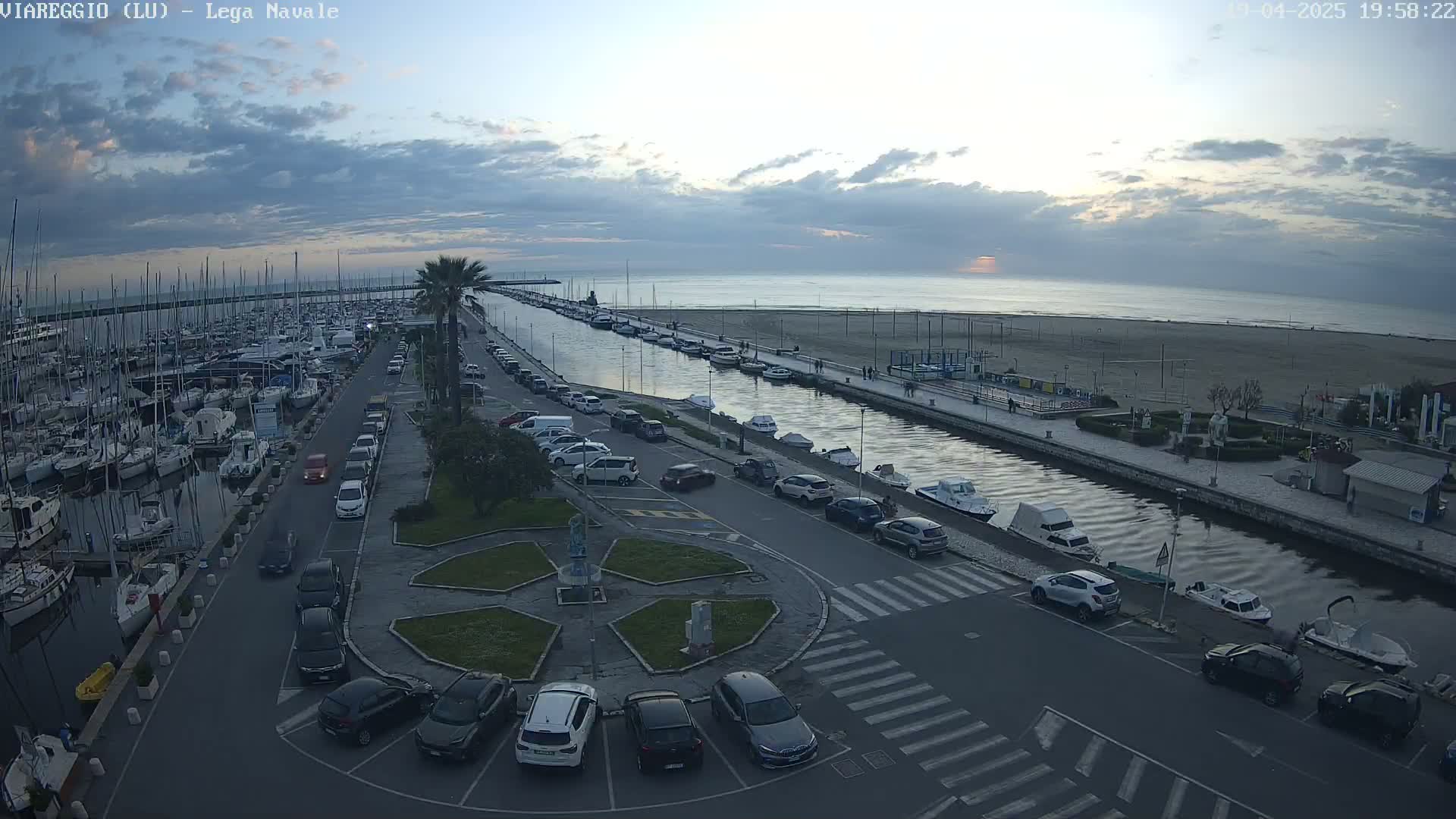 An aerial view of a marina with numerous boats, a canal, and a parking lot filled with cars, all under a partly cloudy sunset sky.