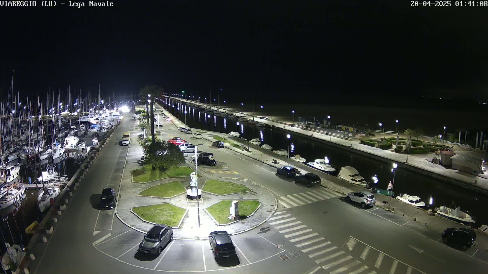 A nighttime aerial view shows a marina filled with boats, a paved area with parked cars, and a canal lined with lights.