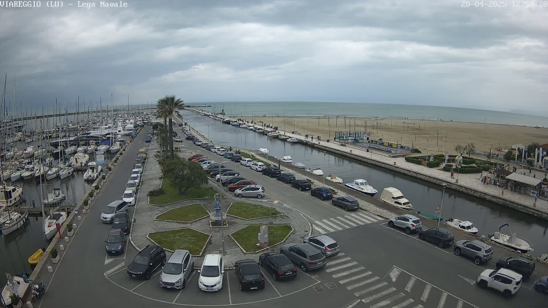 A marina filled with boats is adjacent to a canal lined with parked cars and a sandy beach under a cloudy sky.