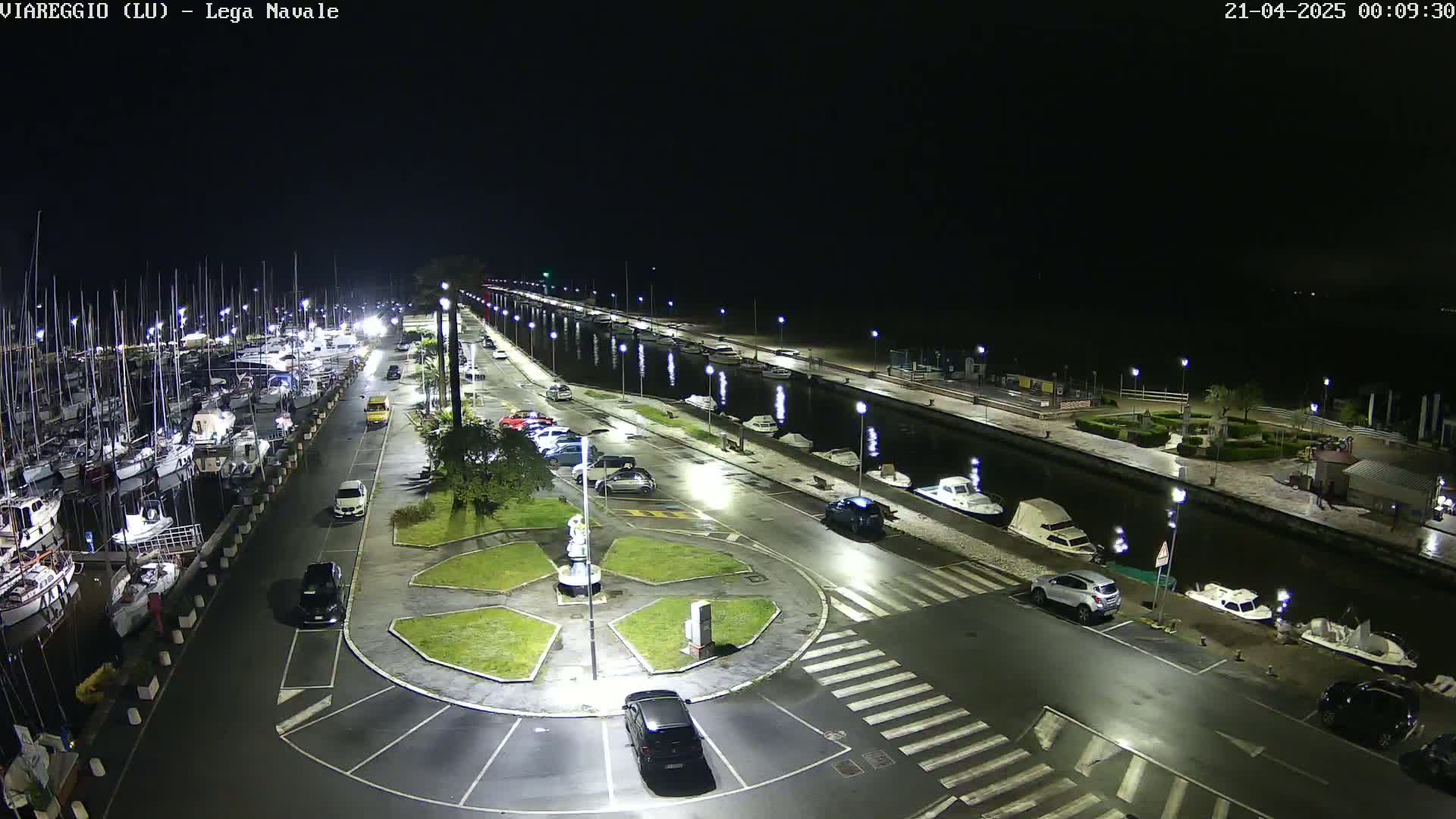 A nighttime aerial view of a marina with numerous boats docked along a canal, surrounded by parked cars and streetlights on a paved area.