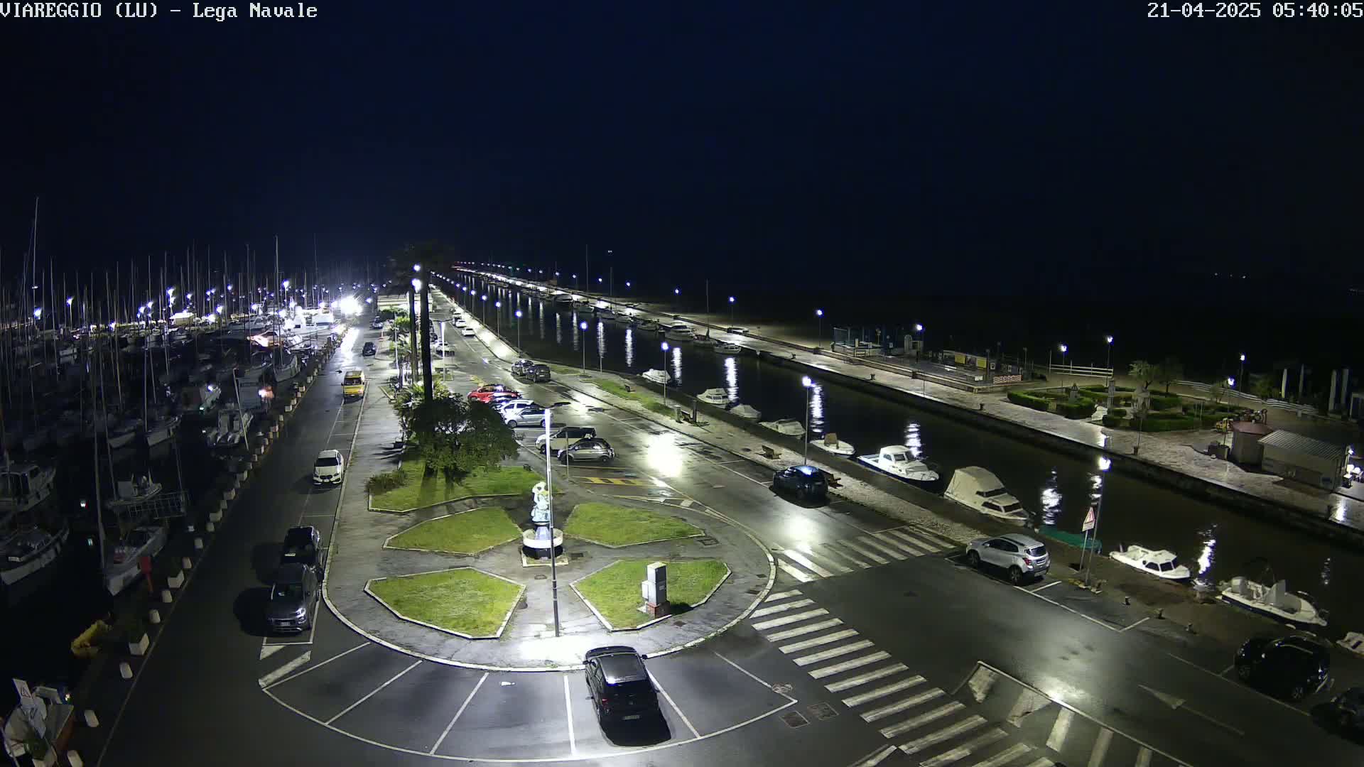 A nighttime aerial view shows a marina filled with boats, a paved area with cars parked, and a canal reflecting lights.