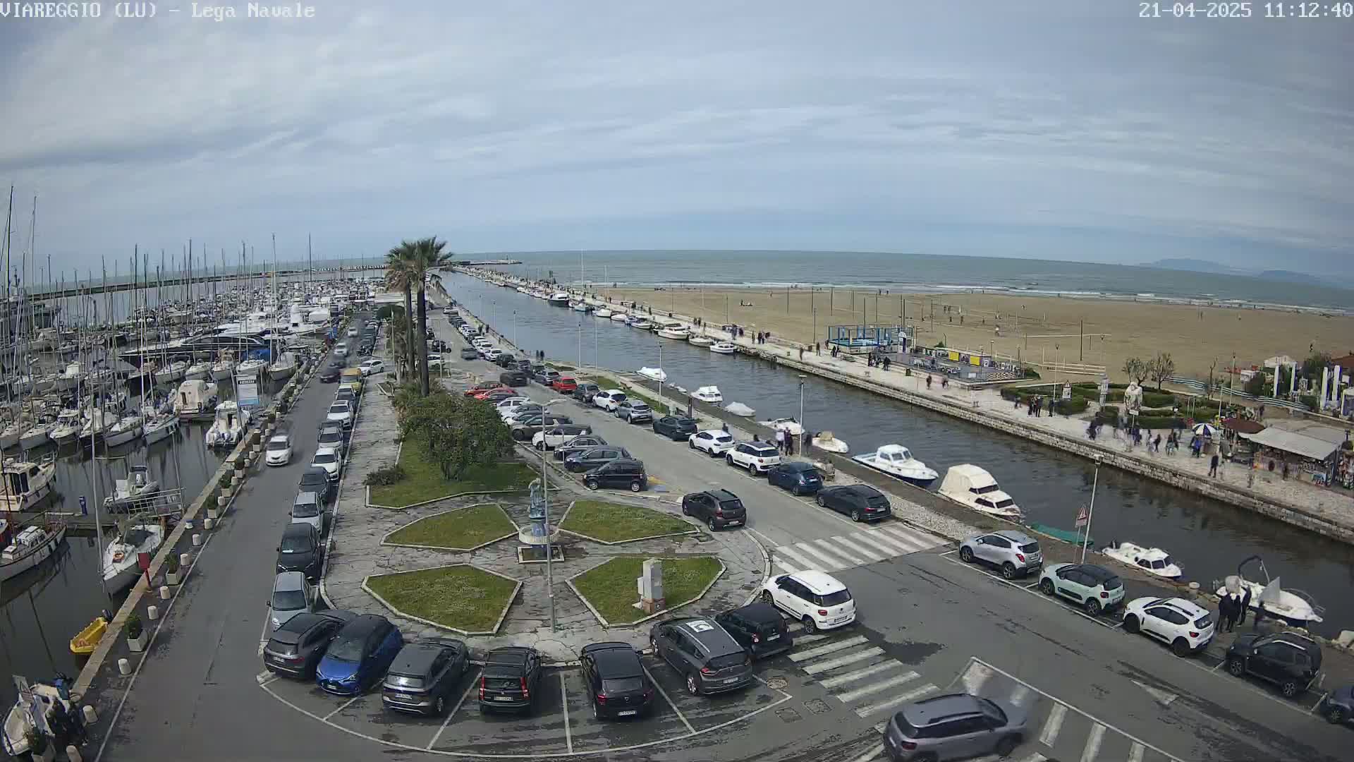 A harbor filled with boats is adjacent to a parking lot and canal that leads to a sandy beach with people walking along the shoreline under a mostly cloudy sky.