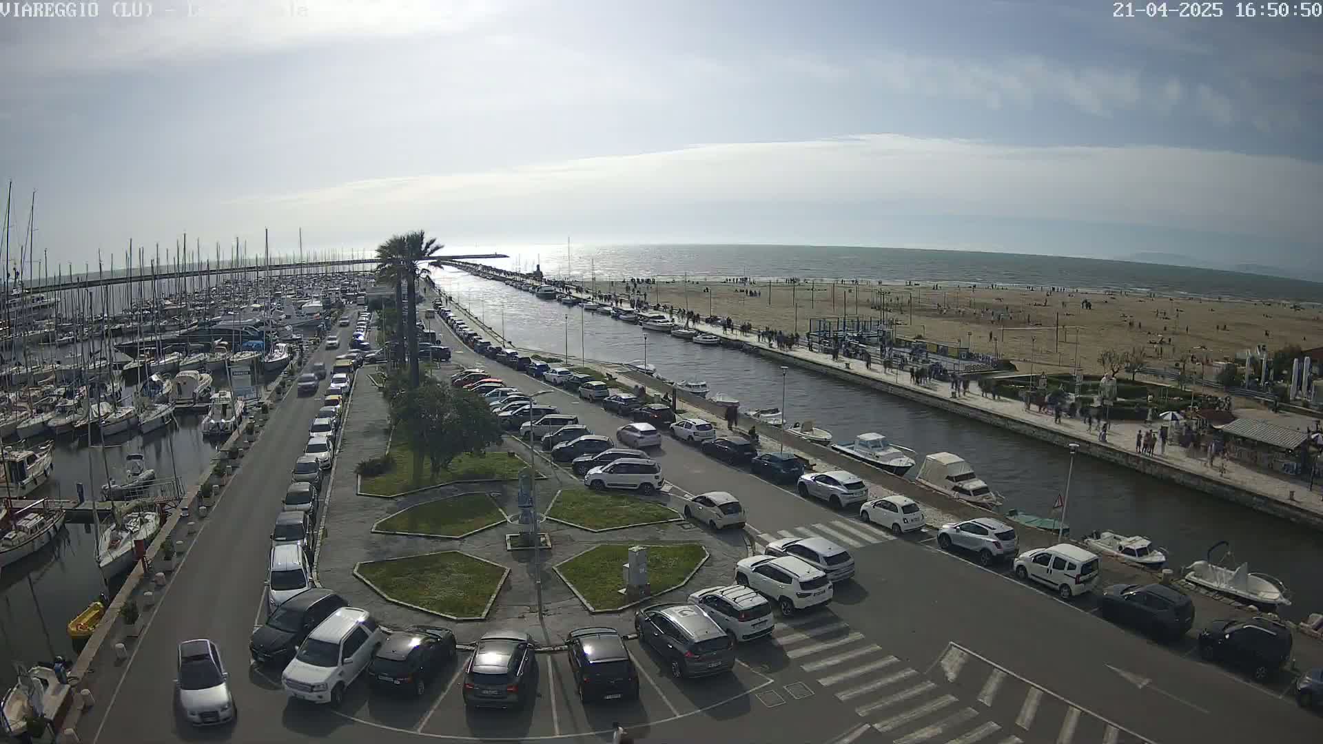 A marina filled with boats is adjacent to a canal and parking lot next to a sandy beach on a partly sunny day.
