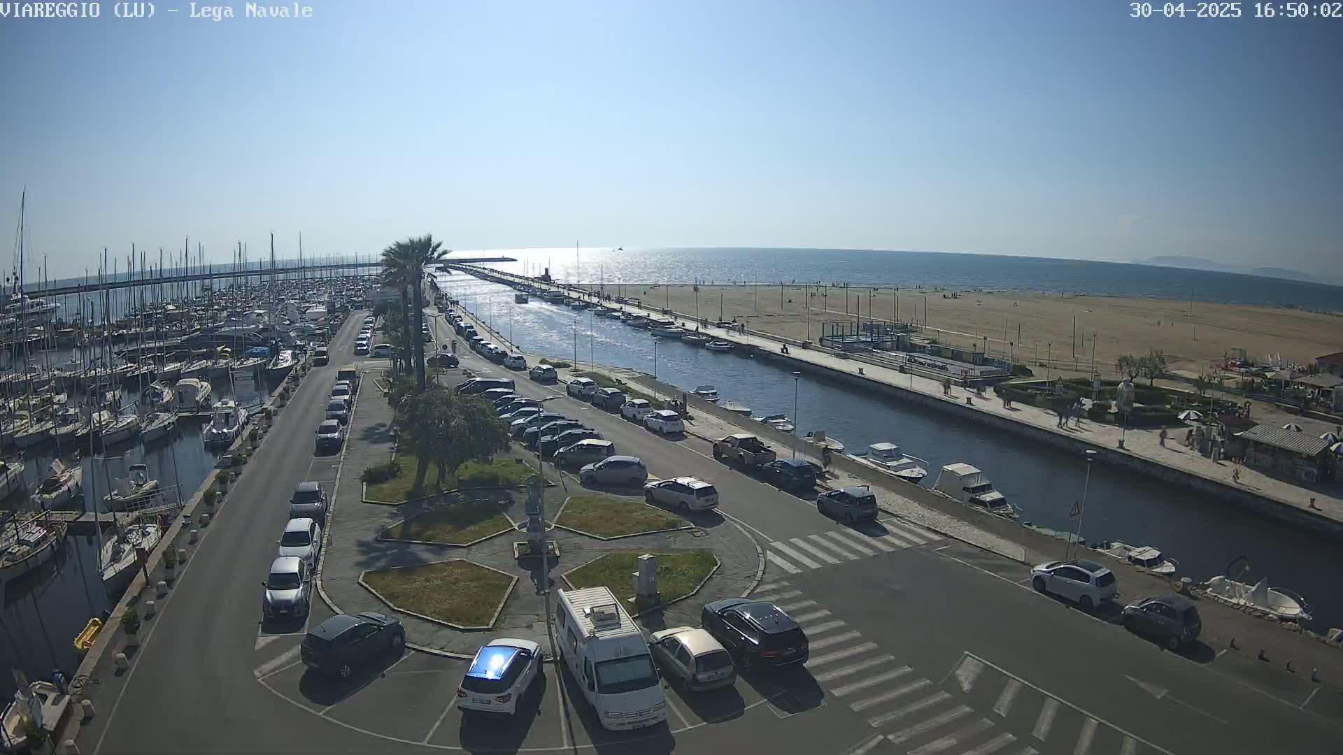 A sunny day at a marina shows a harbor filled with boats, cars parked along a canal-side road, and a sandy beach beyond.