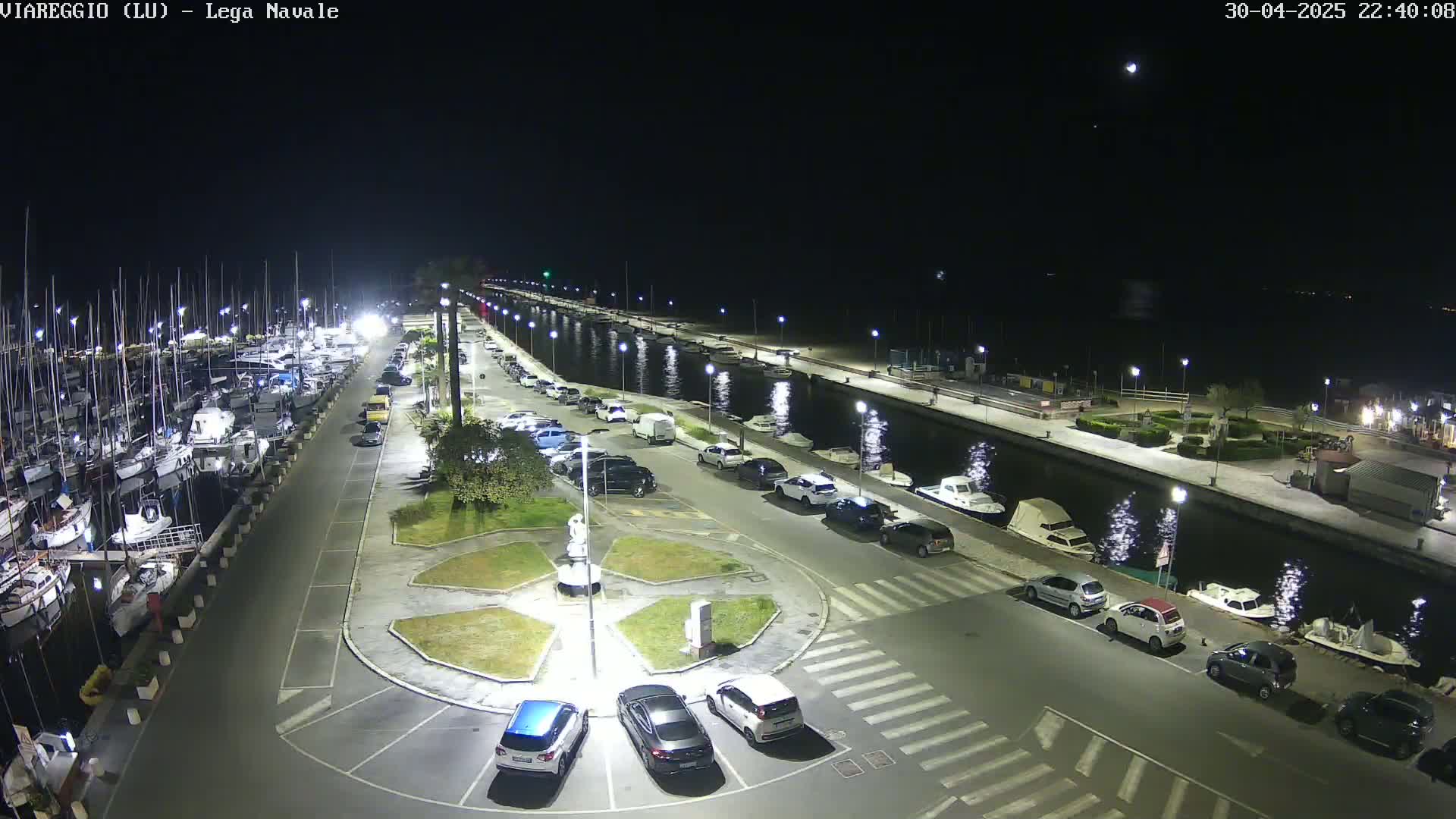 A nighttime view of a harbor with many boats docked, a parking lot filled with cars, and a calm canal reflecting lights.