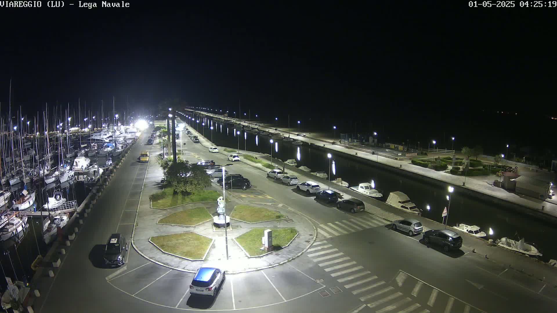 A nighttime aerial view shows a marina filled with boats, a roadway with parked cars, and a circular plaza with a statue in the center, all brightly lit under a clear, dark sky.