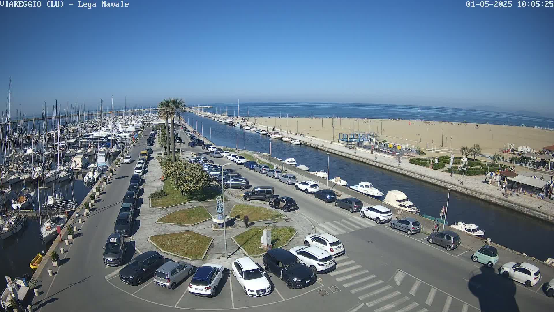 A marina filled with boats is adjacent to a sandy beach with people sunbathing, all under a clear blue sky.