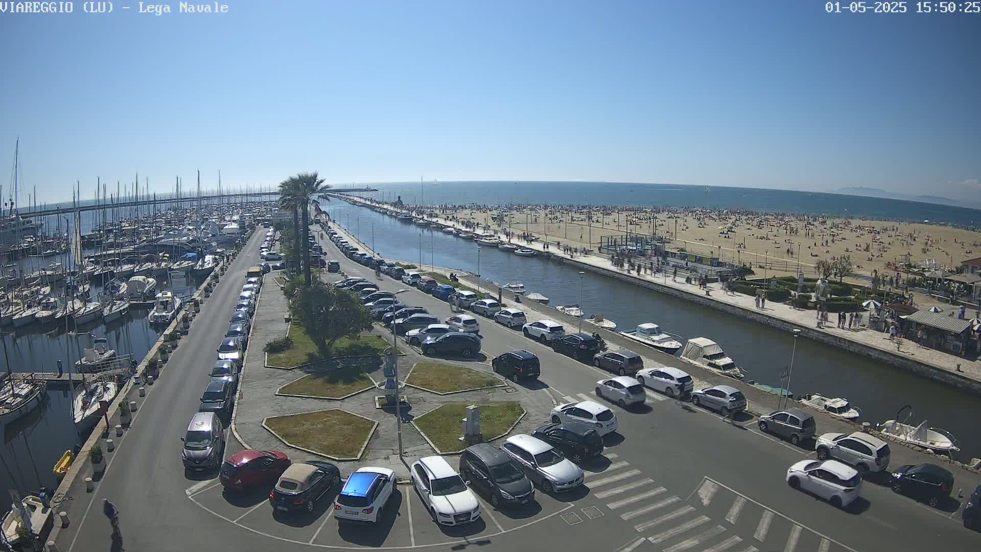 A sunny day at a marina shows a crowded beach beyond a canal lined with parked cars and boats.