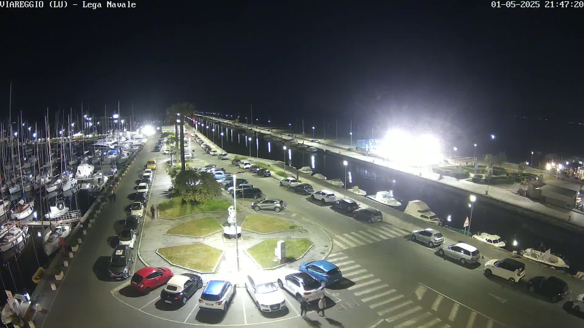 A nighttime, aerial view of a harbor with many boats docked, a parking lot full of cars, and a small plaza in the foreground.