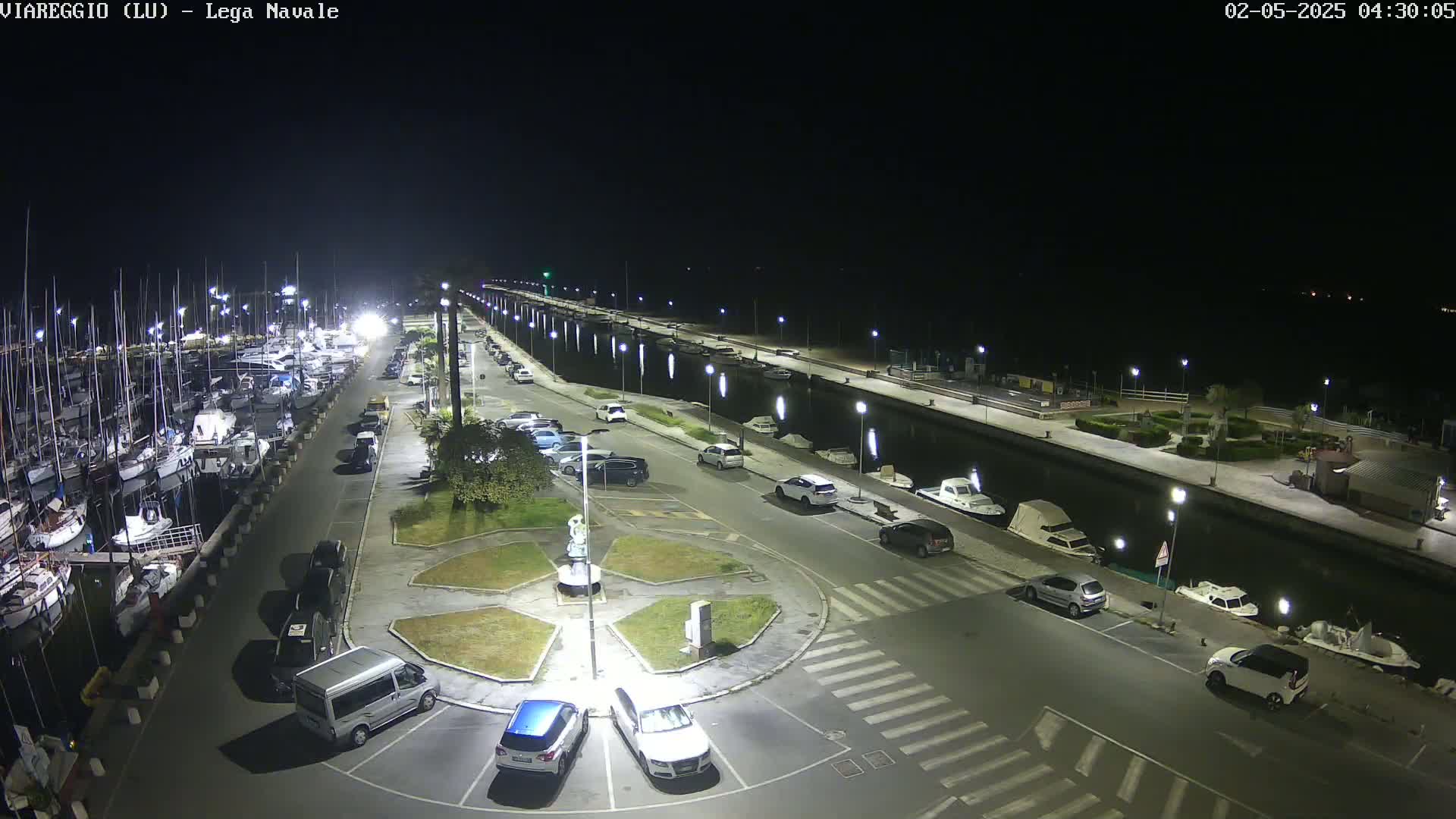 A nighttime aerial view of a marina with numerous boats docked alongside a paved area containing parked cars and a central landscaped roundabout.