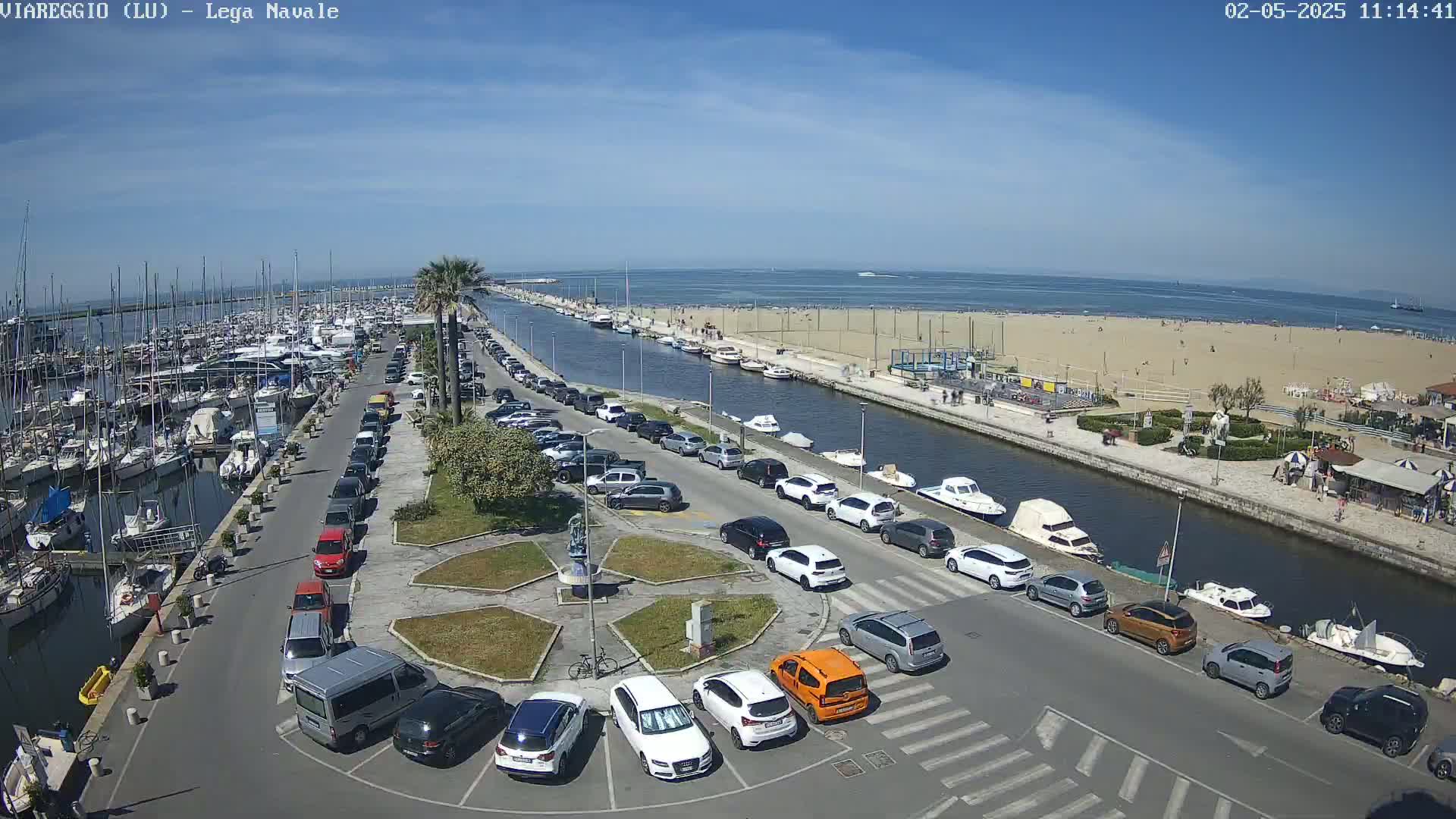 A marina filled with boats, a sandy beach with people, and a parking lot with cars are seen on a sunny day.