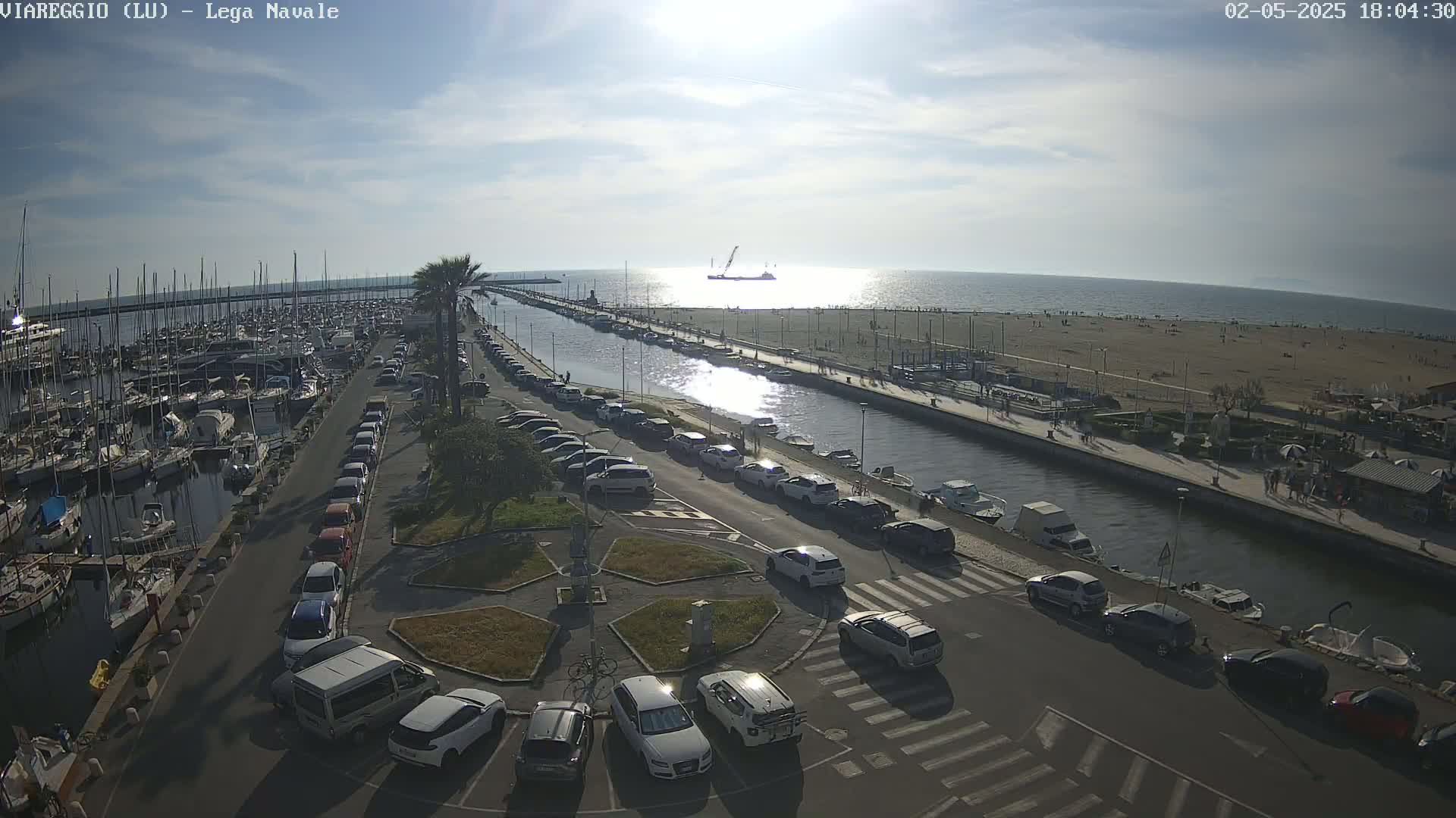 A marina filled with boats is adjacent to a canal, which runs alongside a beach with people on it, all under a partly sunny sky.
