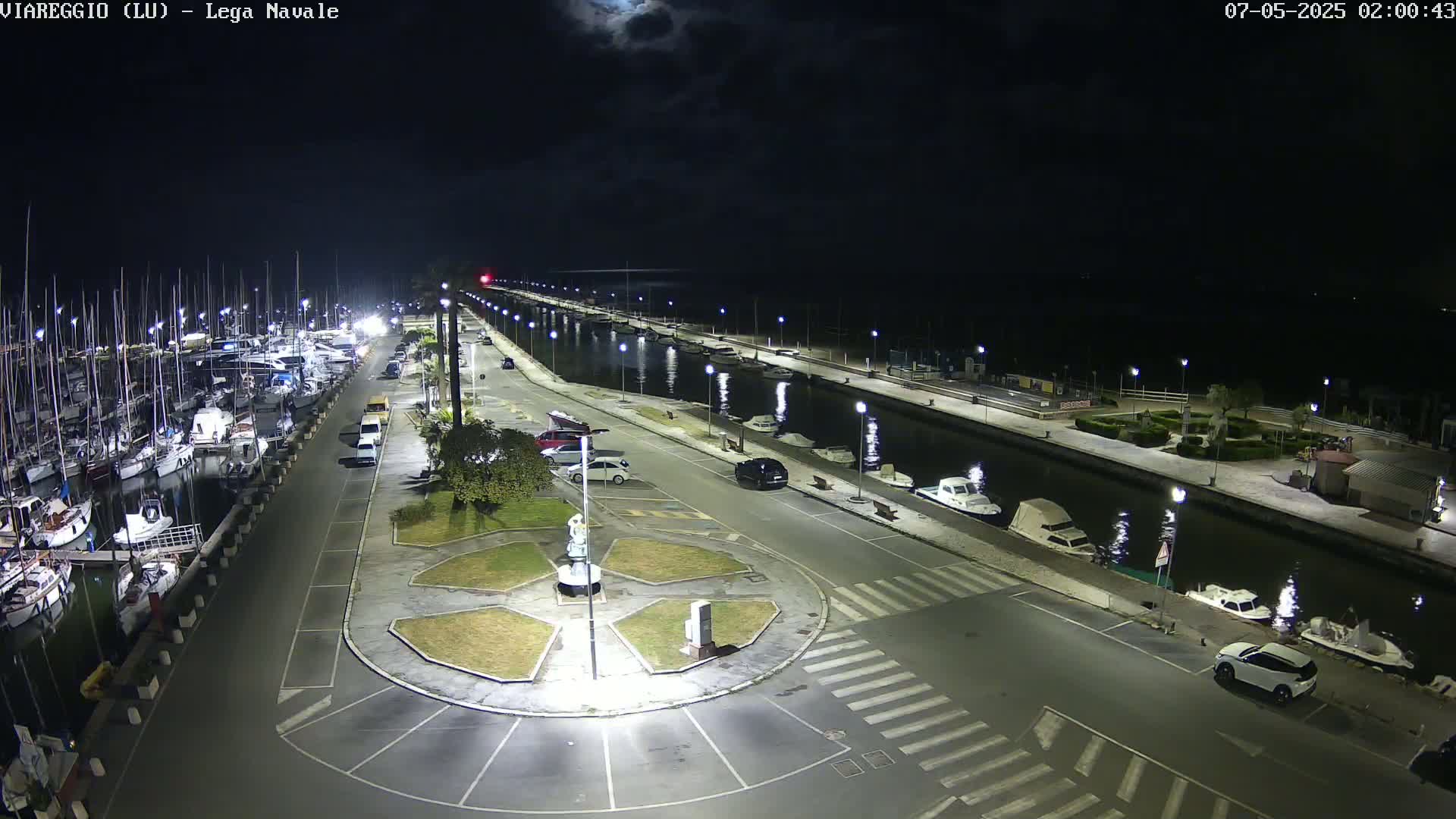 A nighttime, high-angle view of a marina with boats docked along a canal, cars parked in a lot, and a roundabout in the foreground under a mostly clear sky.