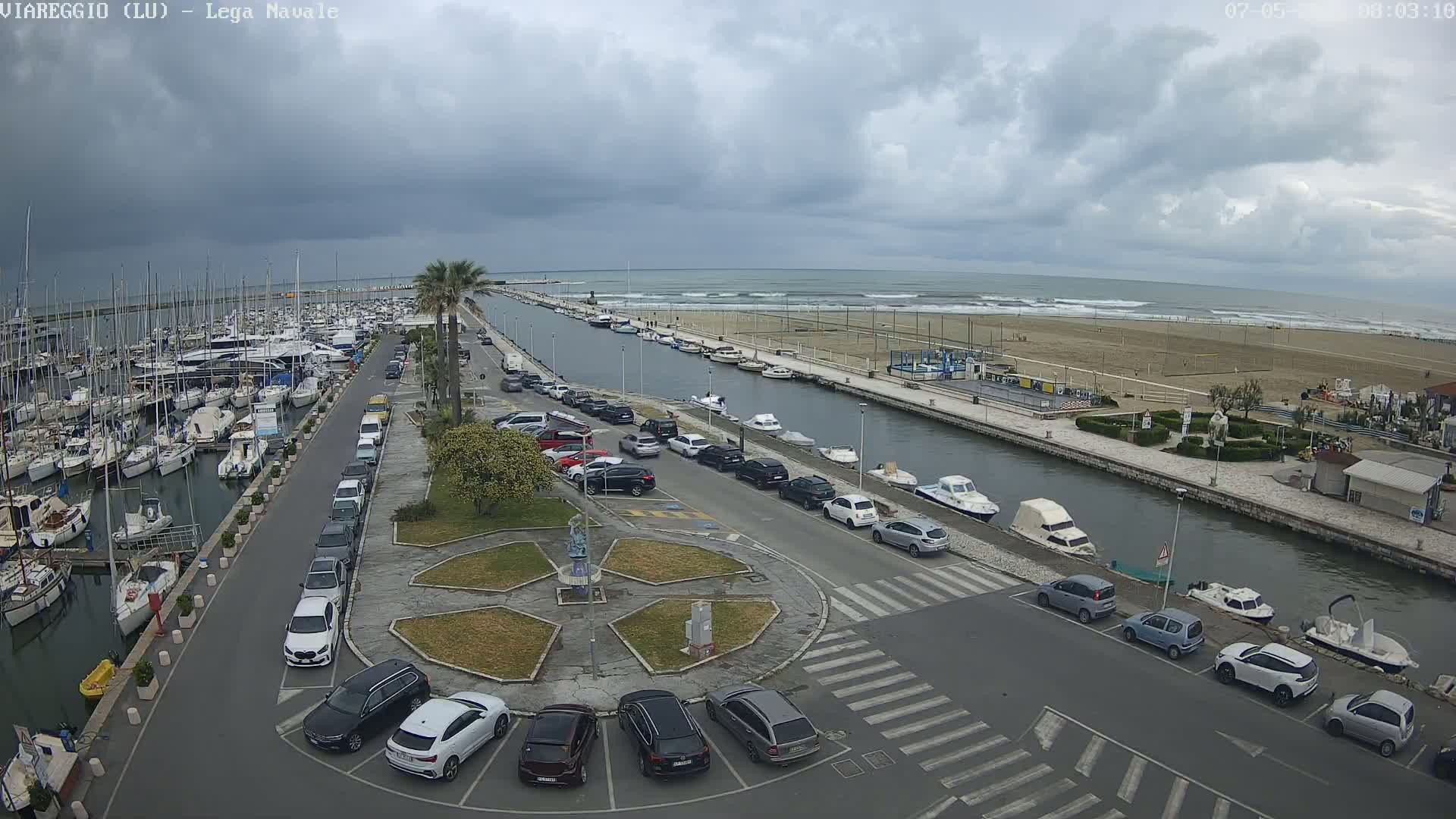 A marina filled with boats is adjacent to a canal that leads to a sandy beach under a cloudy sky.